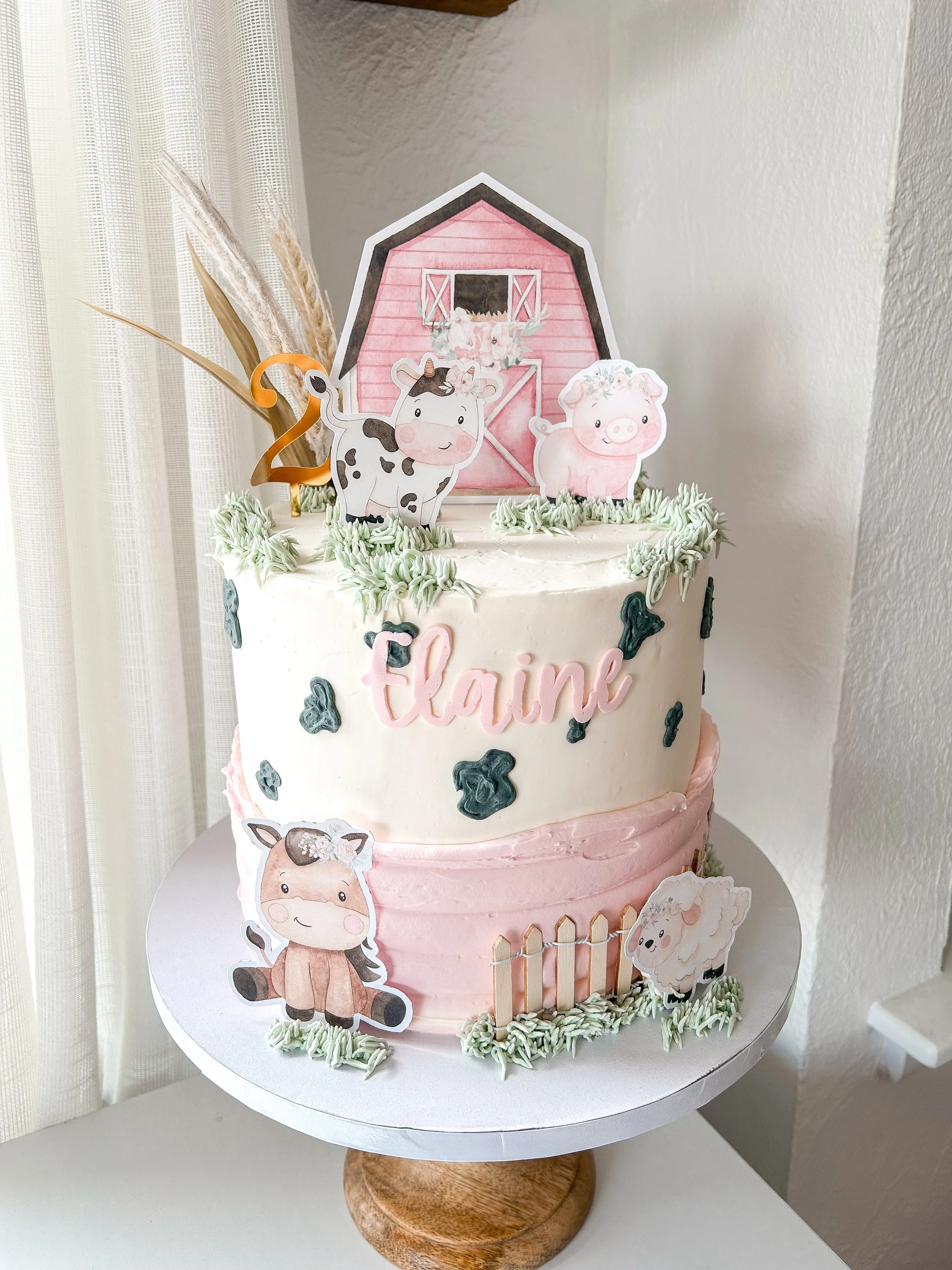 A farm-themed birthday cake decorated with cartoon farm animals, a pink barn topper, and the name 'Elaine' written on the front, set on a white cake stand with a wooden base.