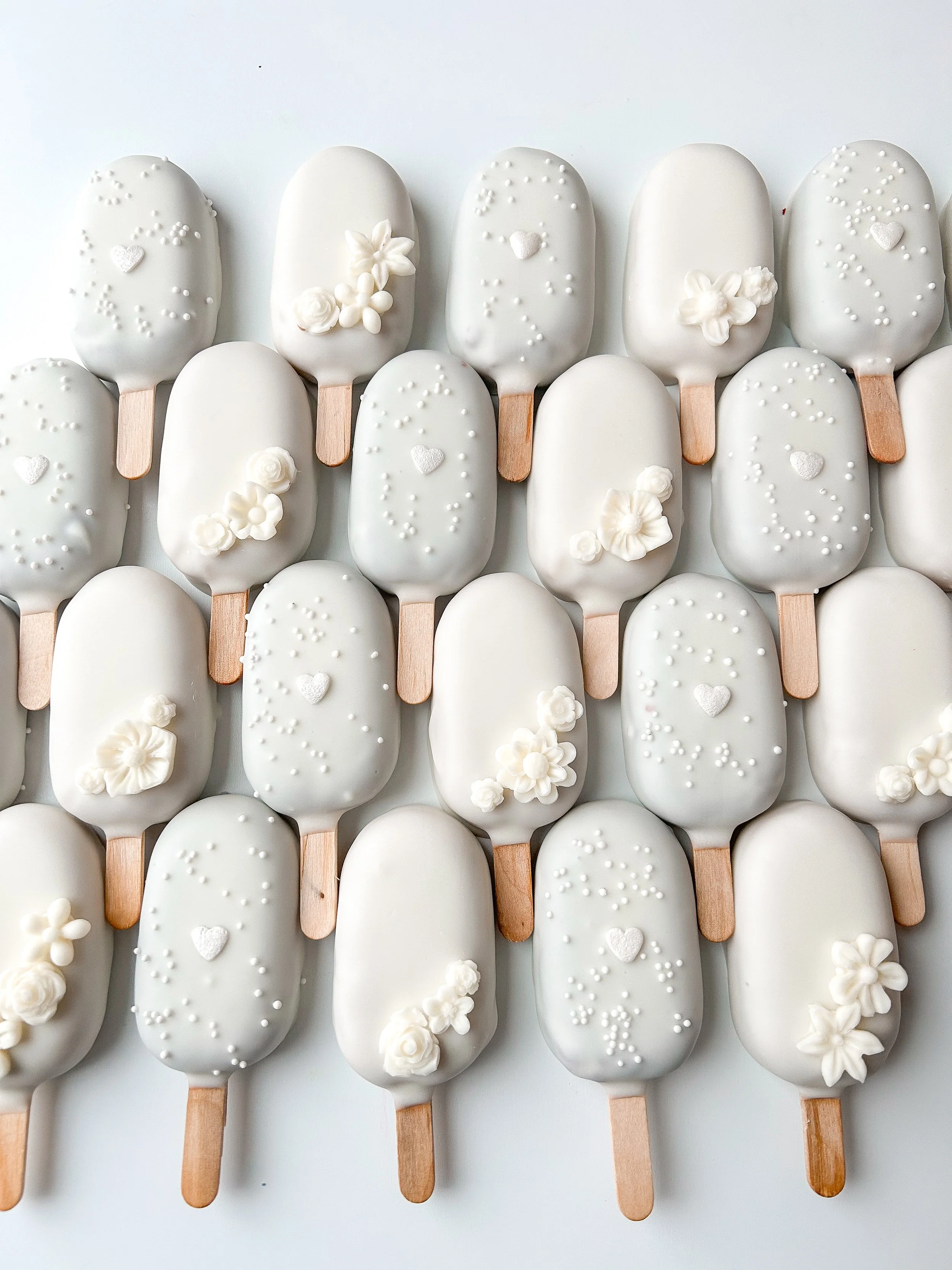 White chocolate-covered ice cream bars decorated with small white flowers, tiny white hearts, and sprinkles, arranged on wooden sticks.
