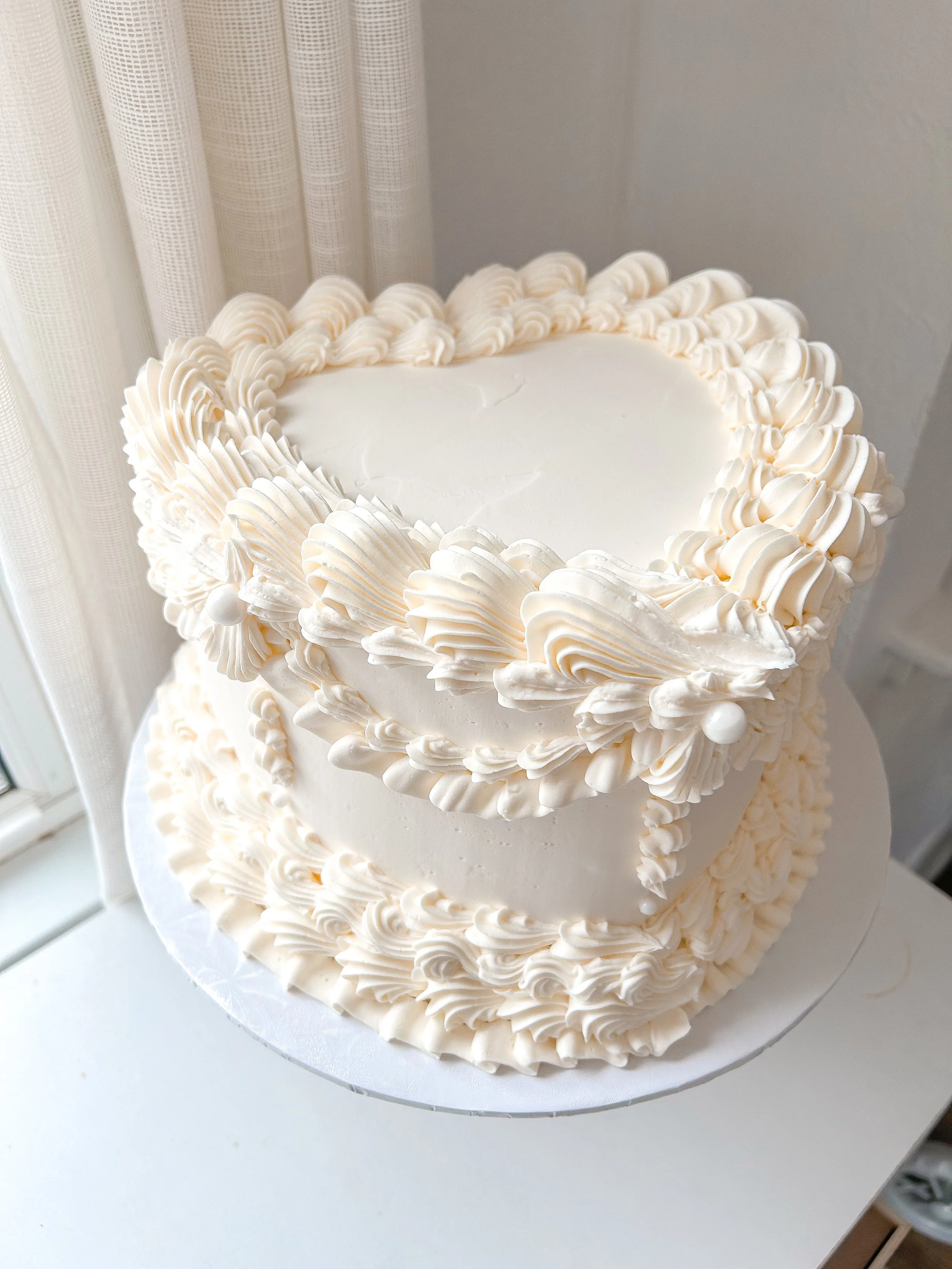 A two-tiered white wedding cake decorated with white frosting rosettes and shell patterns, placed on a white cake board near a window with curtains.