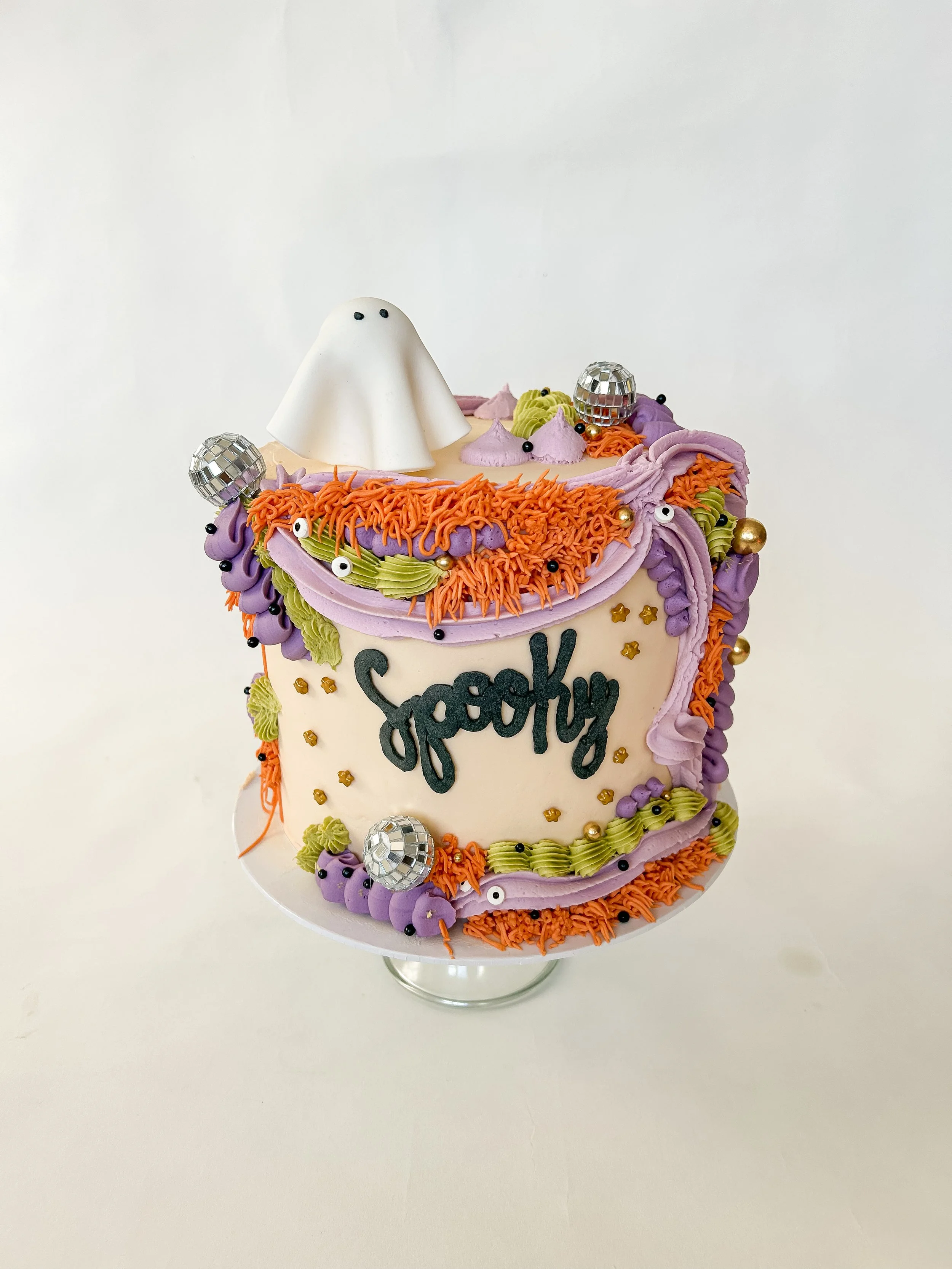 A colorful cake decorated with purple, orange, green, and pink frosting, featuring a ghost figure on top and disco ball decorations, with the name 'Jpoppy' written on the front.