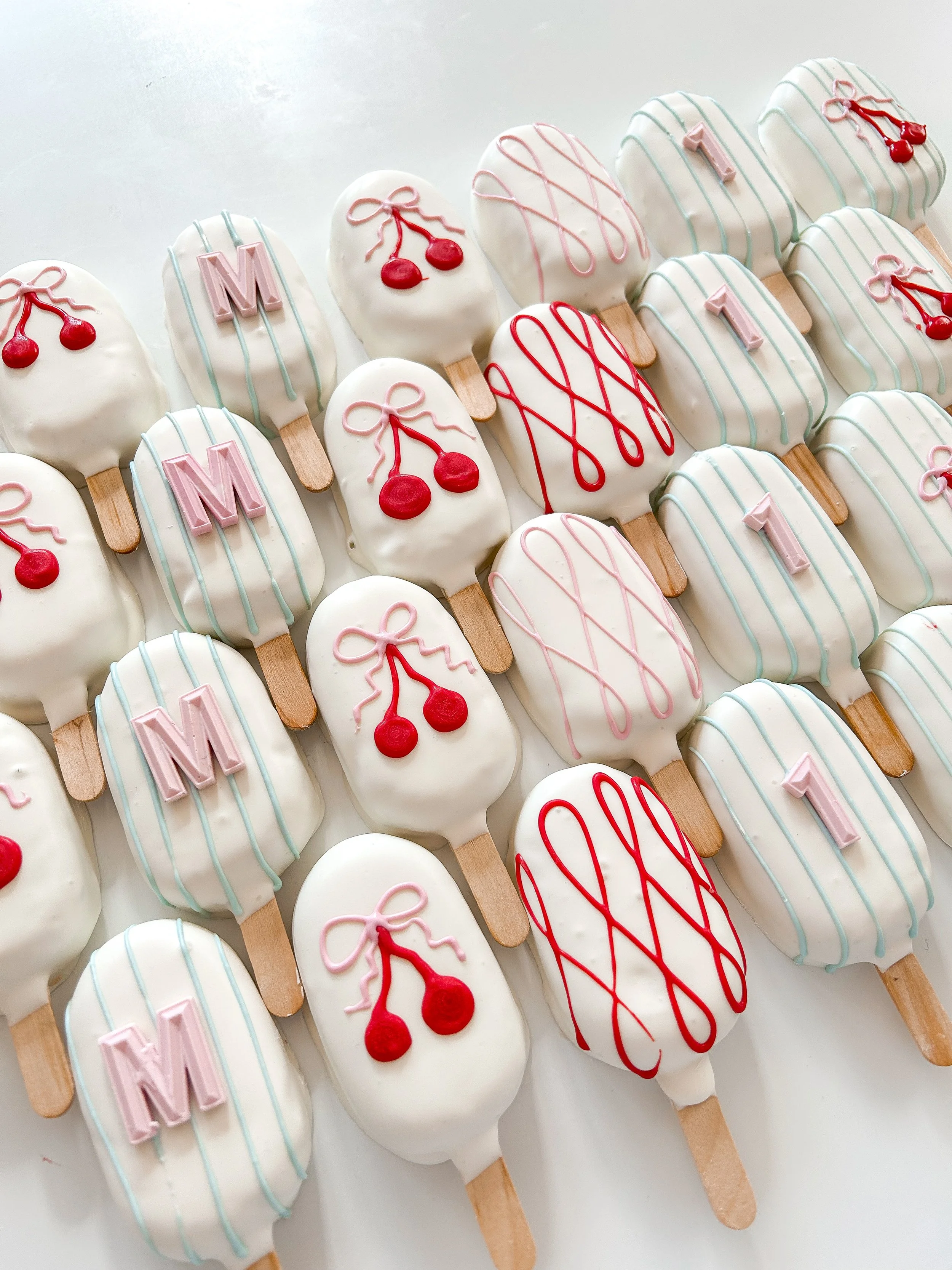 Assorted decorated ice cream bar cookies with cherry and cherry pie decorations, pink and blue drizzle, and letter and number toppers.