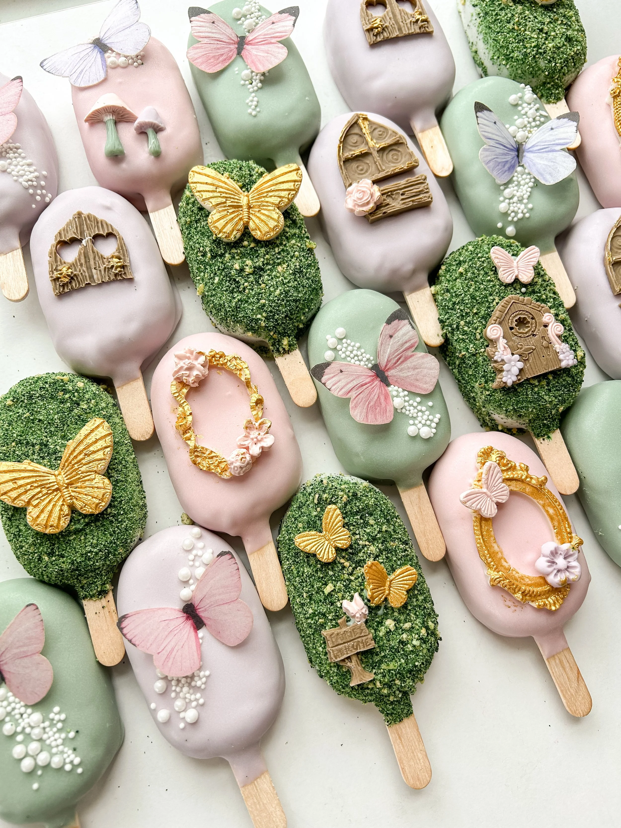 Decorative ice cream bars with pastel pink, green, and white coatings, adorned with butterfly and flower edible decorations, arranged on a white surface.