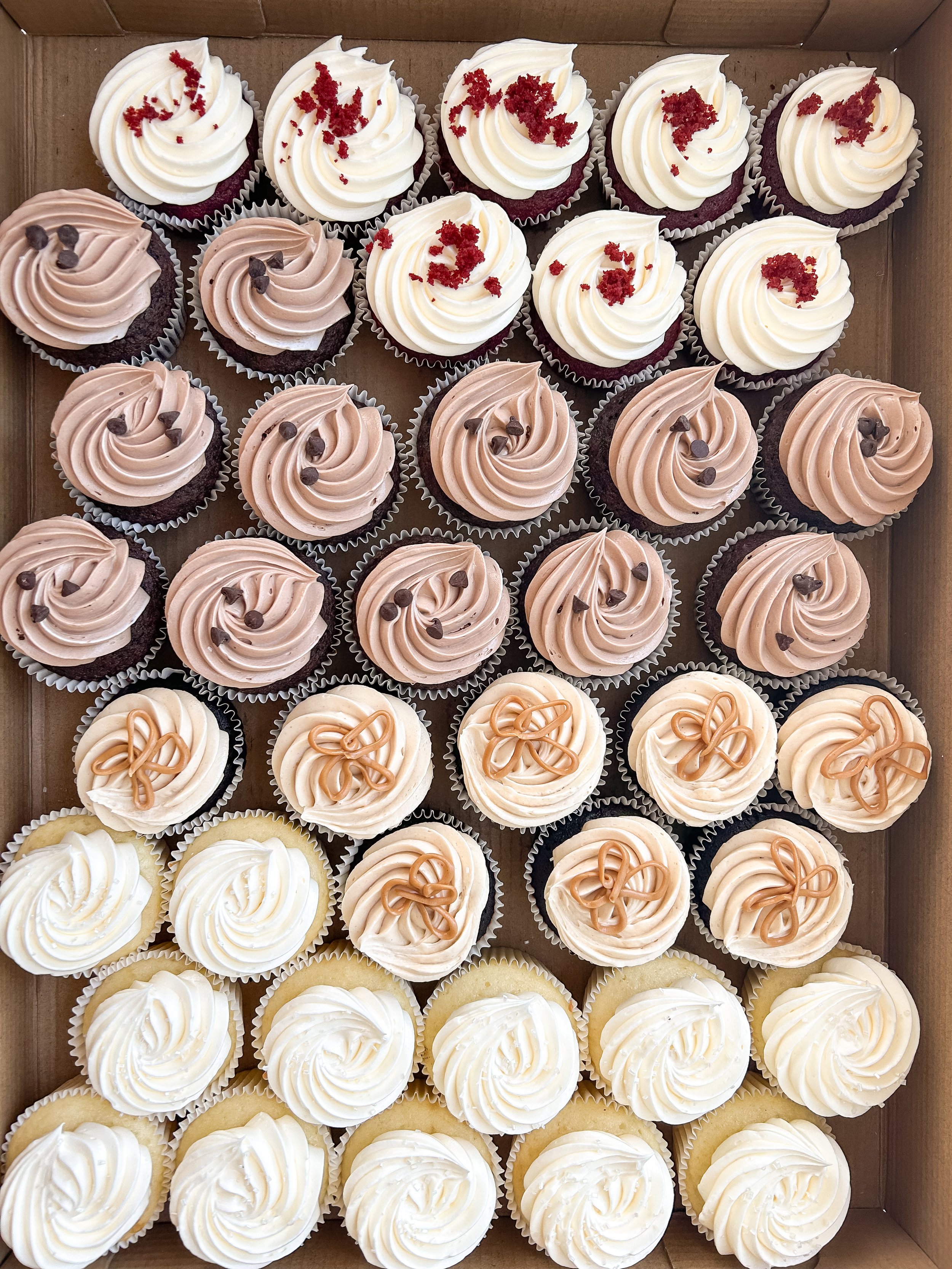 A box of assorted cupcakes with various frosting styles and decorations, including white, chocolate, and caramel-colored frosting.