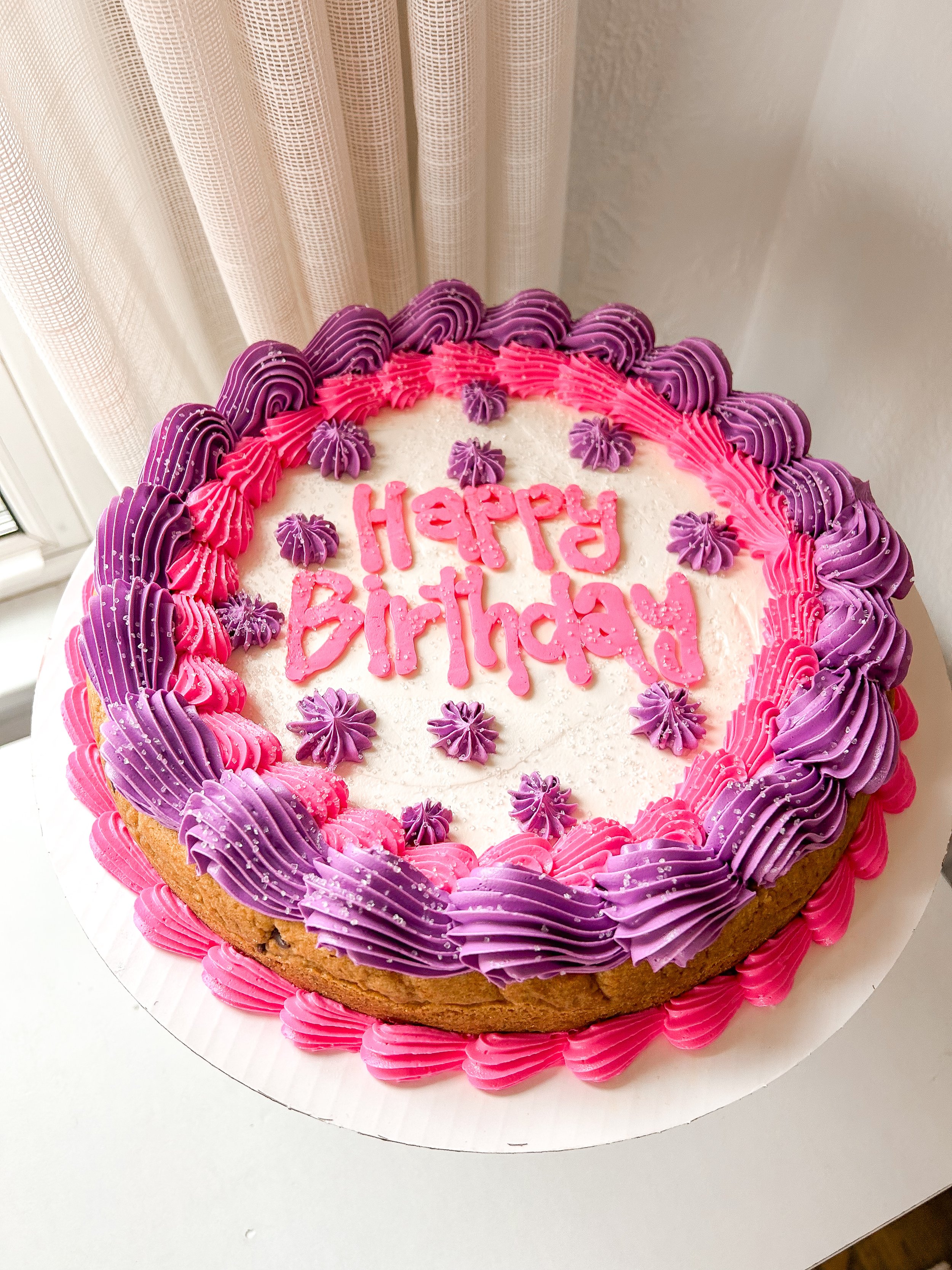 A round birthday cake with pink and purple frosting, decorated with piped rosettes and sprinkles, with 'Happy Birthday' written in pink on top.