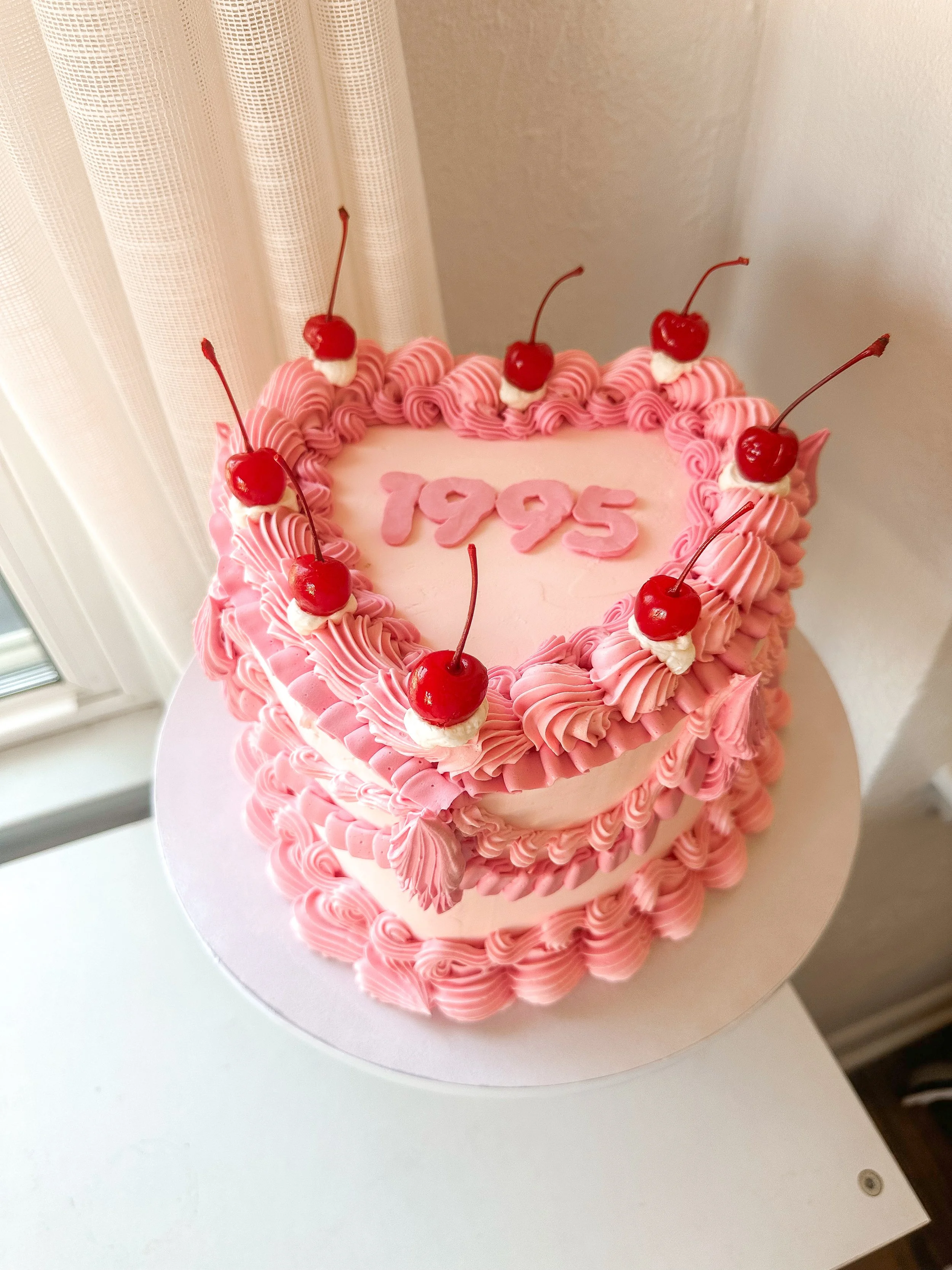 A pink heart-shaped birthday cake with pink and white frosting and cherry toppings, with the year "1995" written in pink on top.