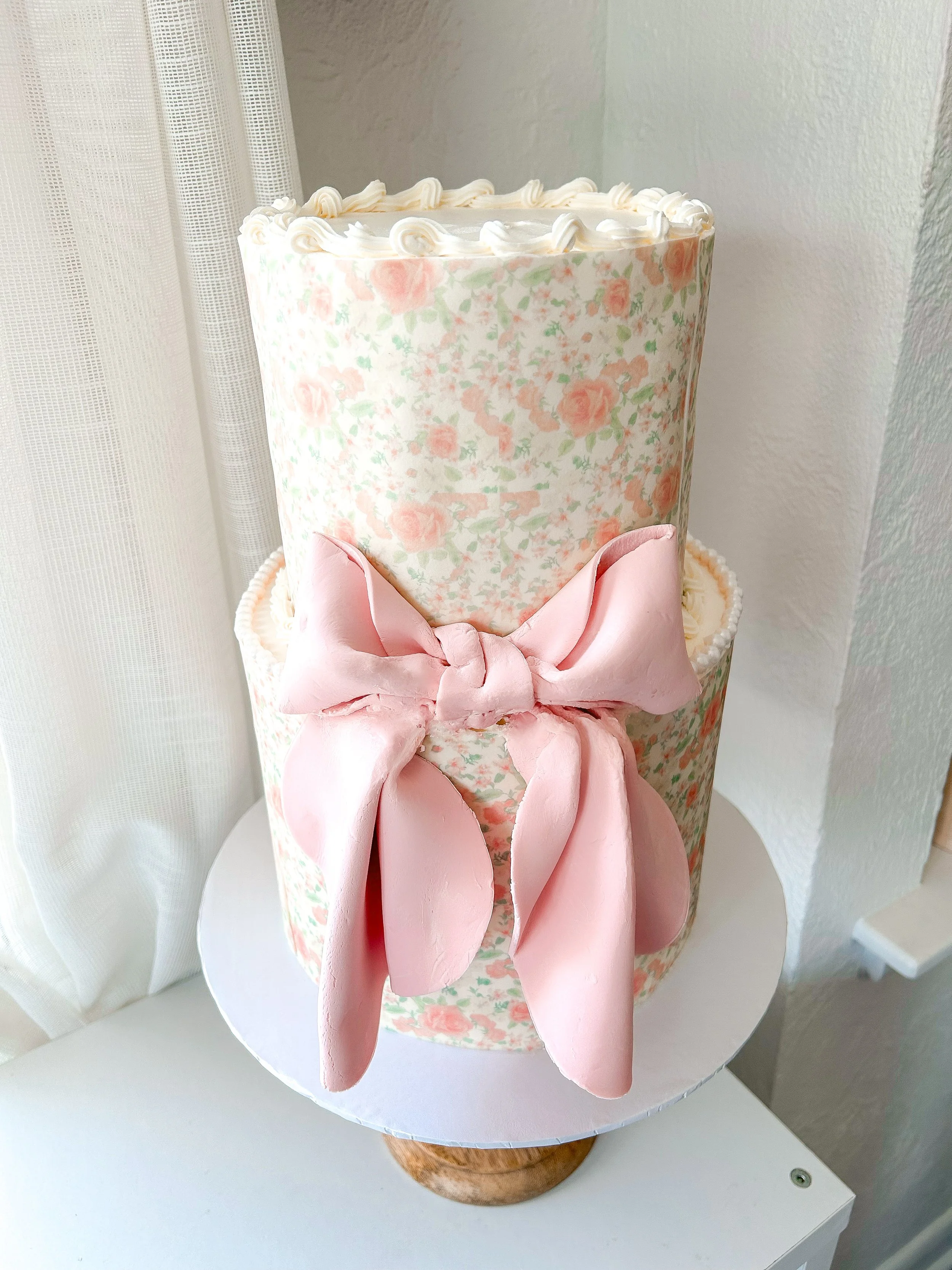A tiered cake decorated with pink floral paper and a large pink fondant bow, placed on a white cake stand in front of white curtains.