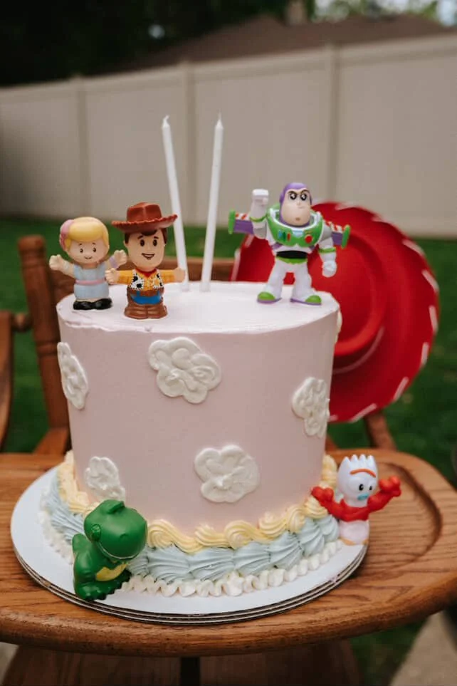 A Toy Story-themed birthday cake with four character figurines: Woody, Buzz Lightyear, Rex, and Forky, decorated with white clouds and pastel-colored icing.
