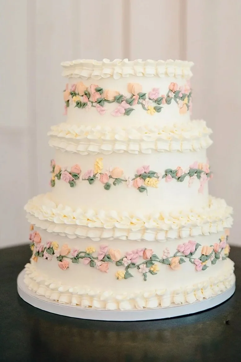 A tall, four-tiered white wedding cake decorated with pink, yellow, and green floral designs and piped white icing borders.