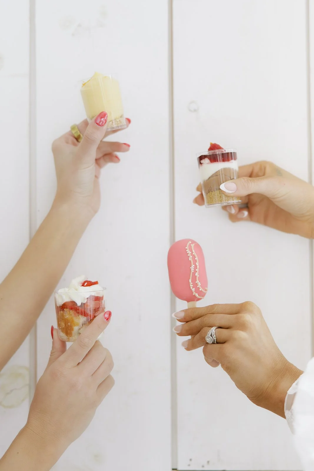 Four hands holding various desserts, including cups with strawberries and cream, a yellow layered cake, a pink decorated popsicle, and a small jar with a layered dessert topped with strawberries and a white sauce.