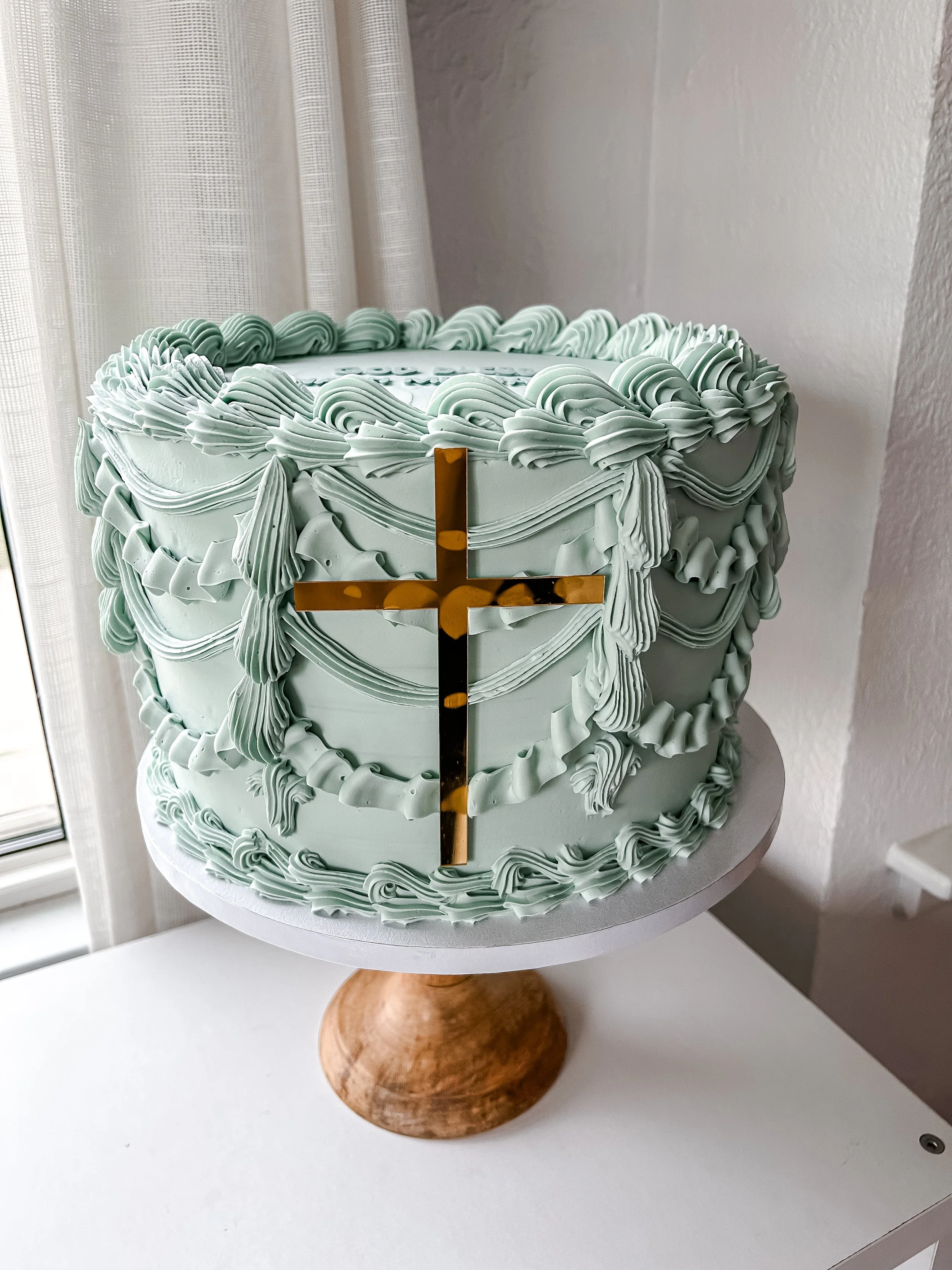 A round, light green cake decorated with piped cream swirls and ropes, featuring a gold cross on the front, placed on a white cake stand with a wooden base near a window with curtains.