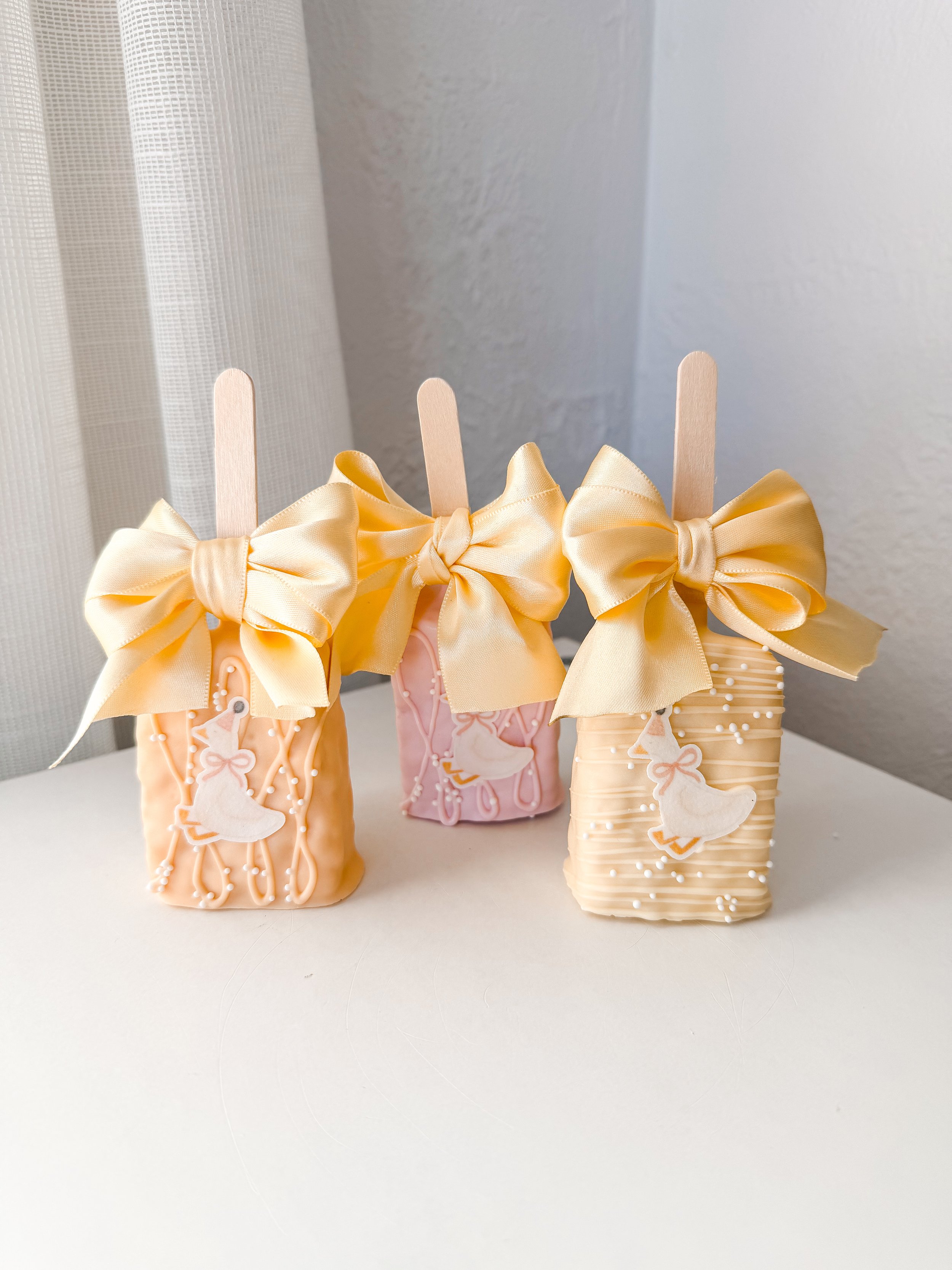 Three decorated cookies with duck and ballet slipper designs, each wrapped with a yellow satin bow on sticks.