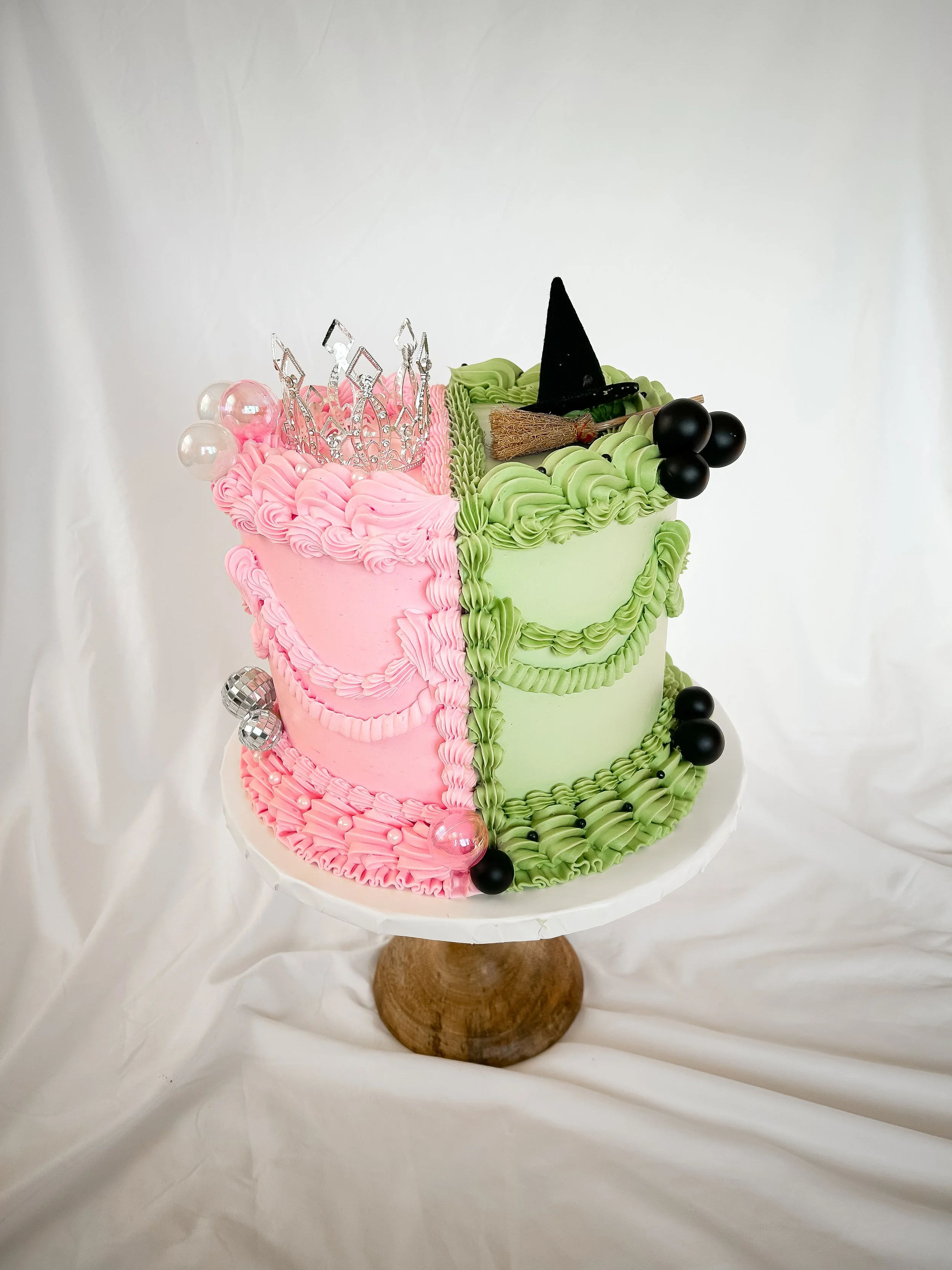 Two-tiered birthday cake with pink on the left and green on the right, decorated with piped frosting, a pink tiara, a black witch hat, black and silver ball decorations, and placed on a white stand with a wooden base.