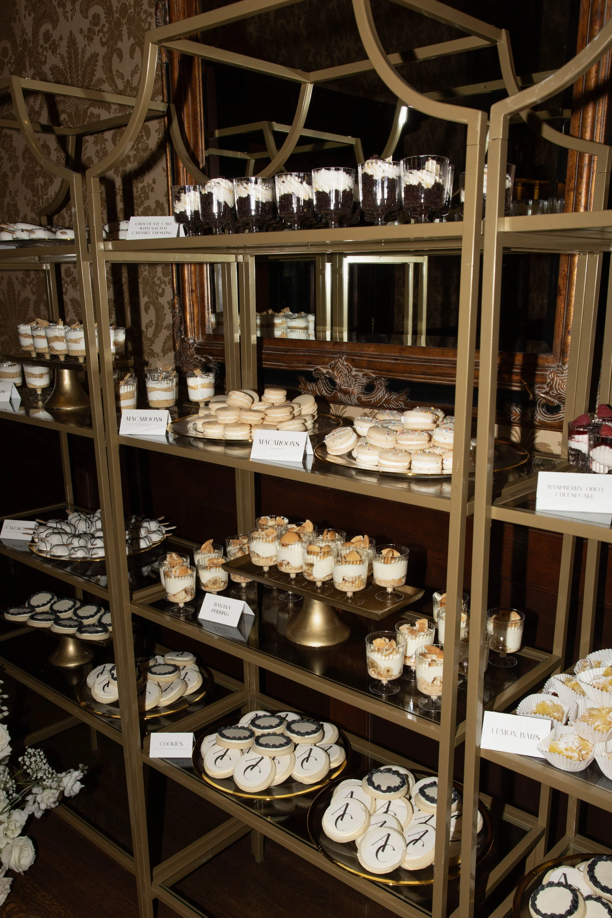 Dessert tray with various cakes, macarons, cookies, lemon bars, and layered parfaits on a gold and glass shelving unit in an elegant setting.