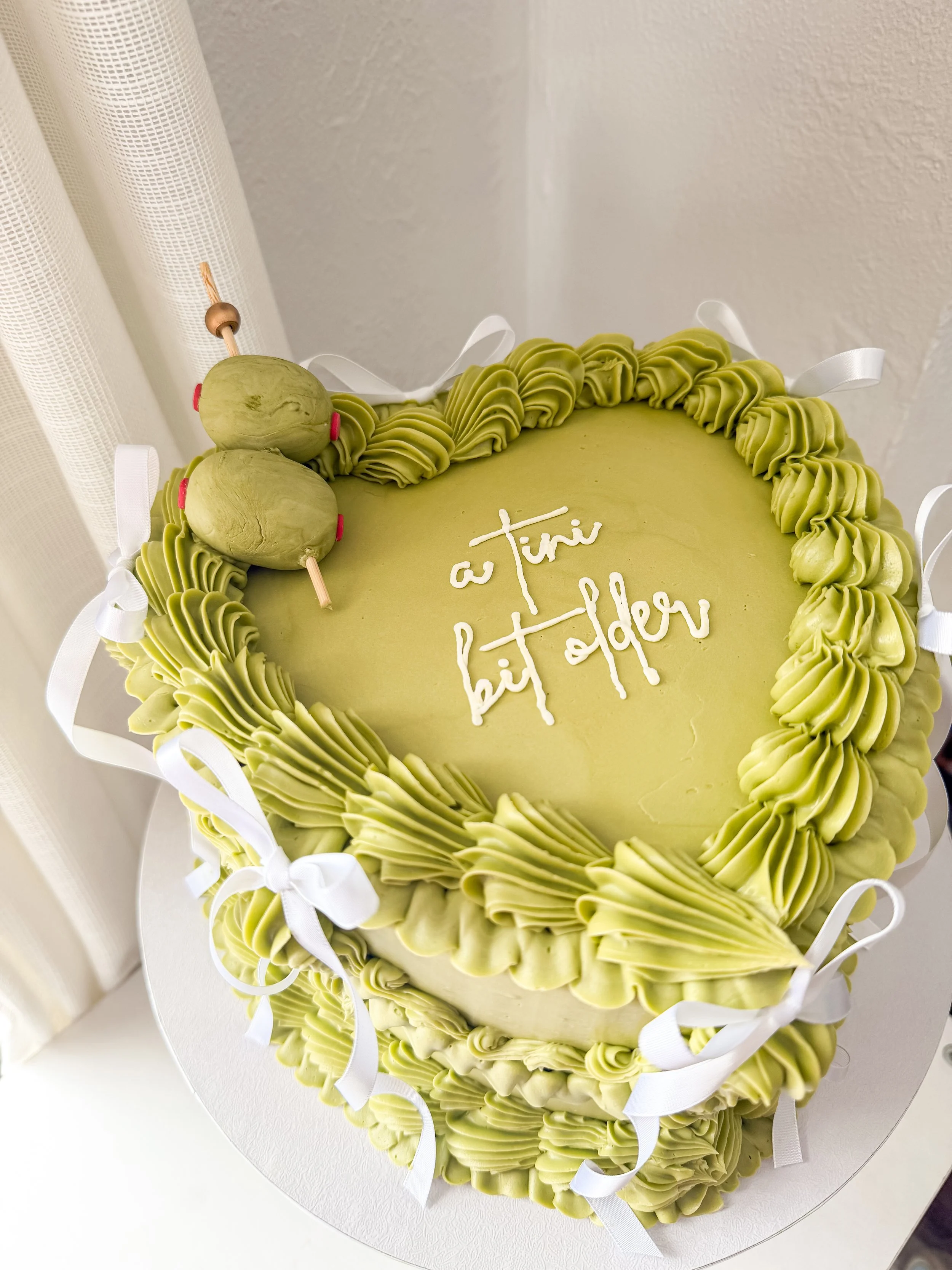 A heart-shaped cake with green frosting, decorated with white ribbons and two green cake pops on a stick, with the words "a tiny bit older" written on top.