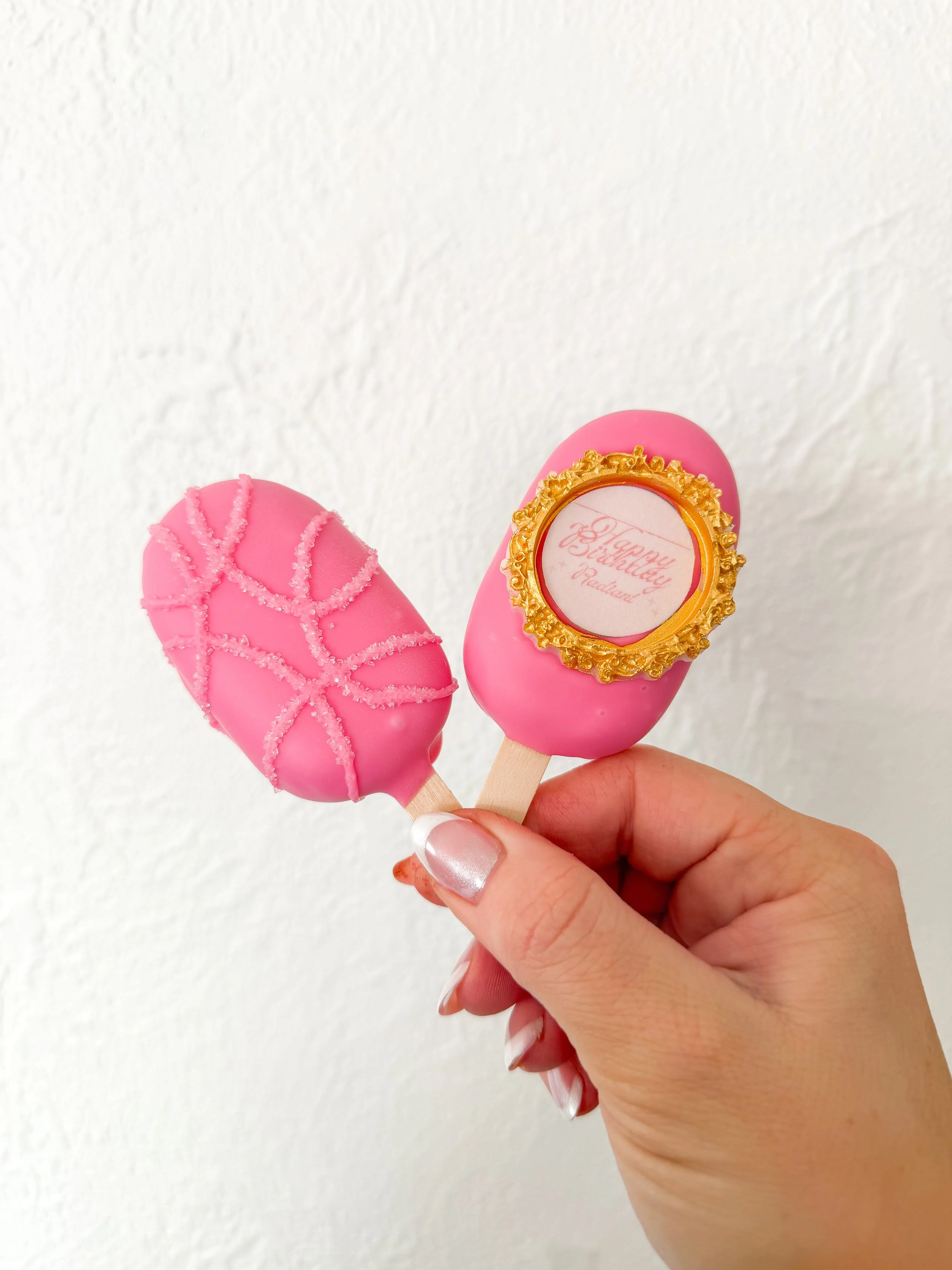 Two pink cake pops, one decorated with white sugar web and the other with a gold ornate frame and a message that reads, 'Happy Eyelash' on a white background.