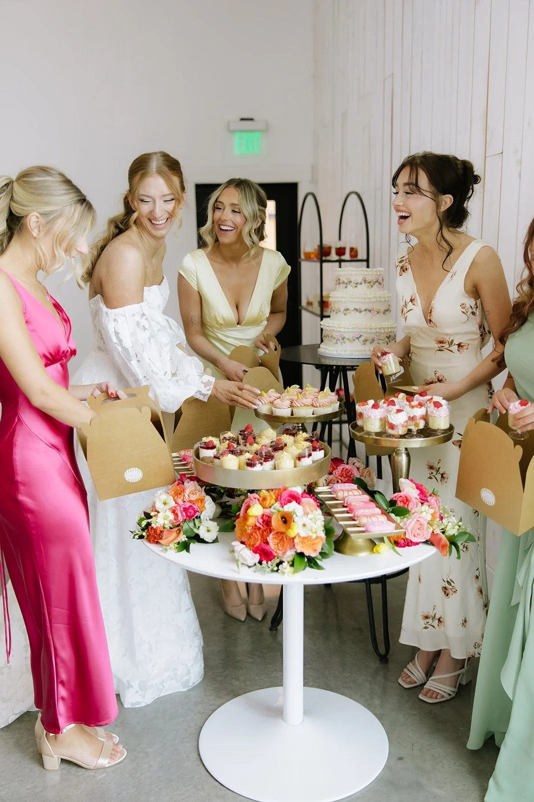 Women in colorful dresses celebrating around a table with desserts and flowers at a bridal shower.