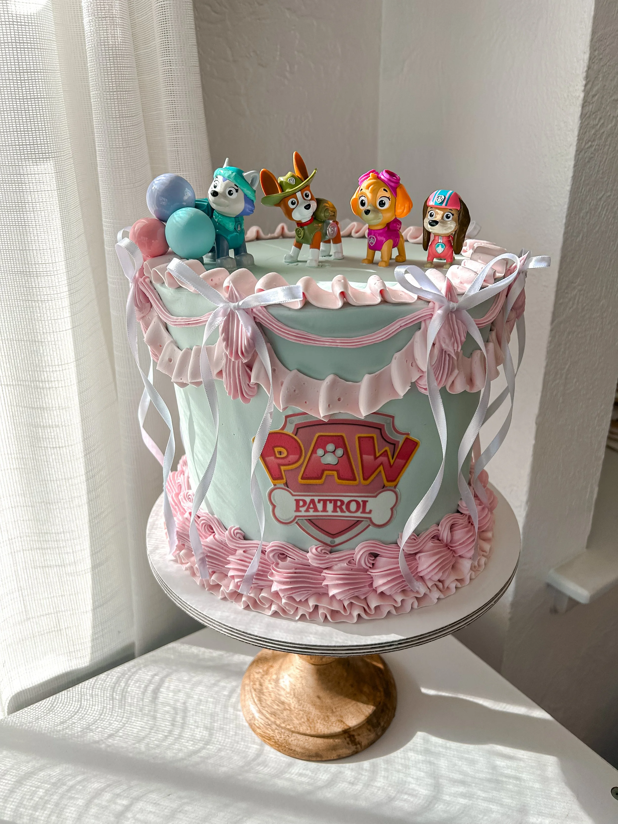 A Paw Patrol themed birthday cake decorated with pink and white icing, featuring four Paw Patrol character figurines on top and colorful balloons.