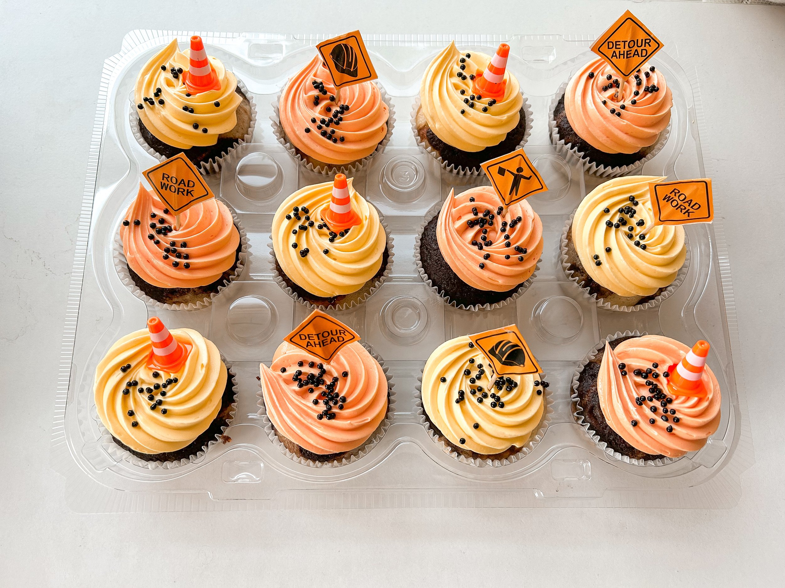 A plastic tray holding ten cupcakes decorated with pink and yellow icing, small black sprinkles, traffic cone toppers, and orange warning signs with messages like "Road Work," "Detour Ahead," and traffic symbols.