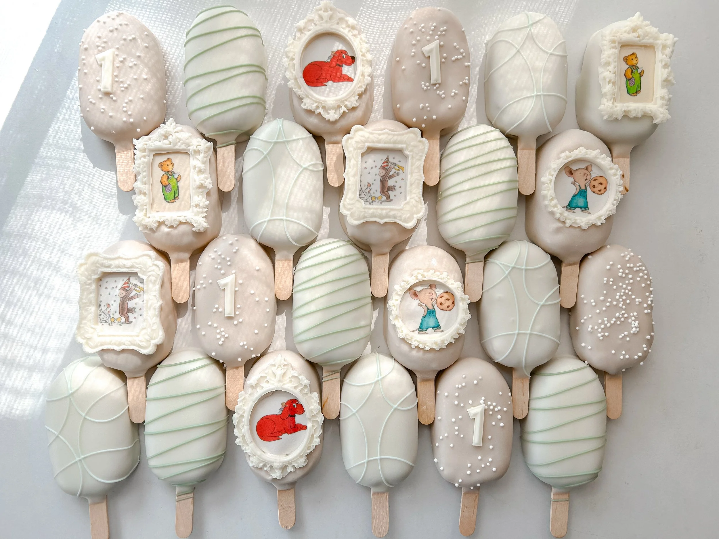 A collection of decorated cookies and cake pops with various designs, including animals, numbers, and cartoon characters, arranged on a white surface.