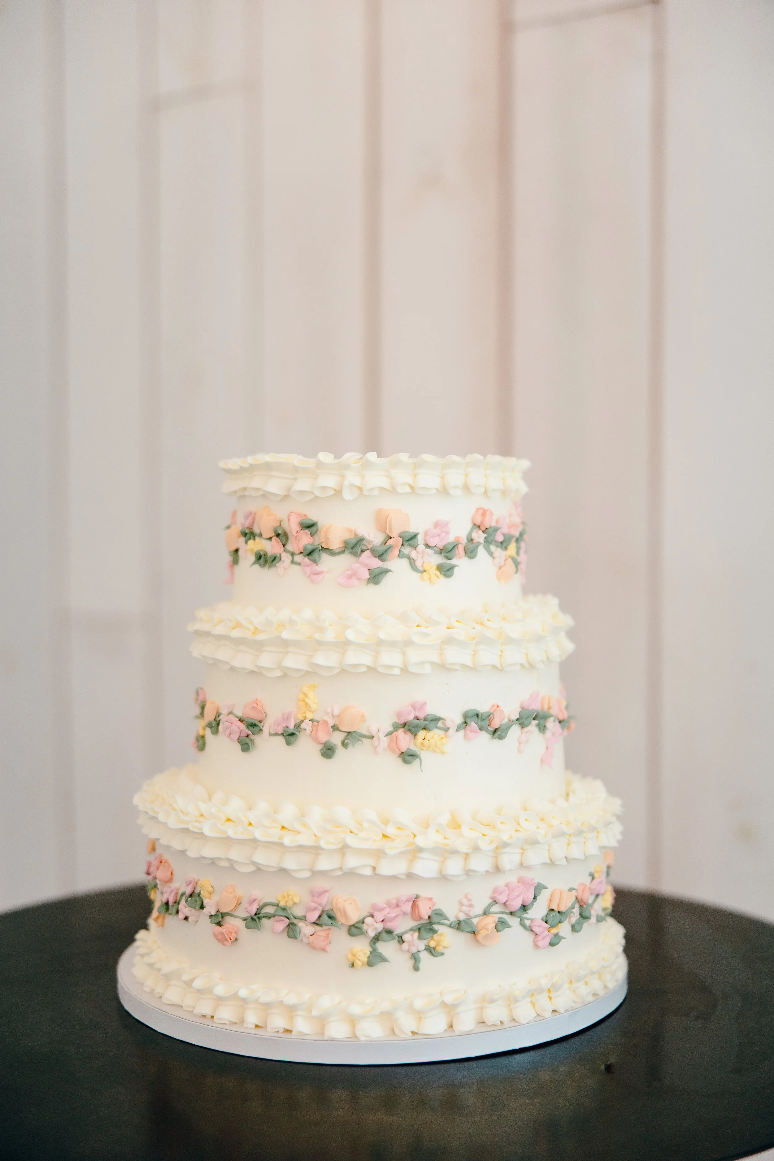 A four-tiered white wedding cake decorated with pastel-colored floral icing and piped white borders, placed on a black surface against a light-colored wooden wall.