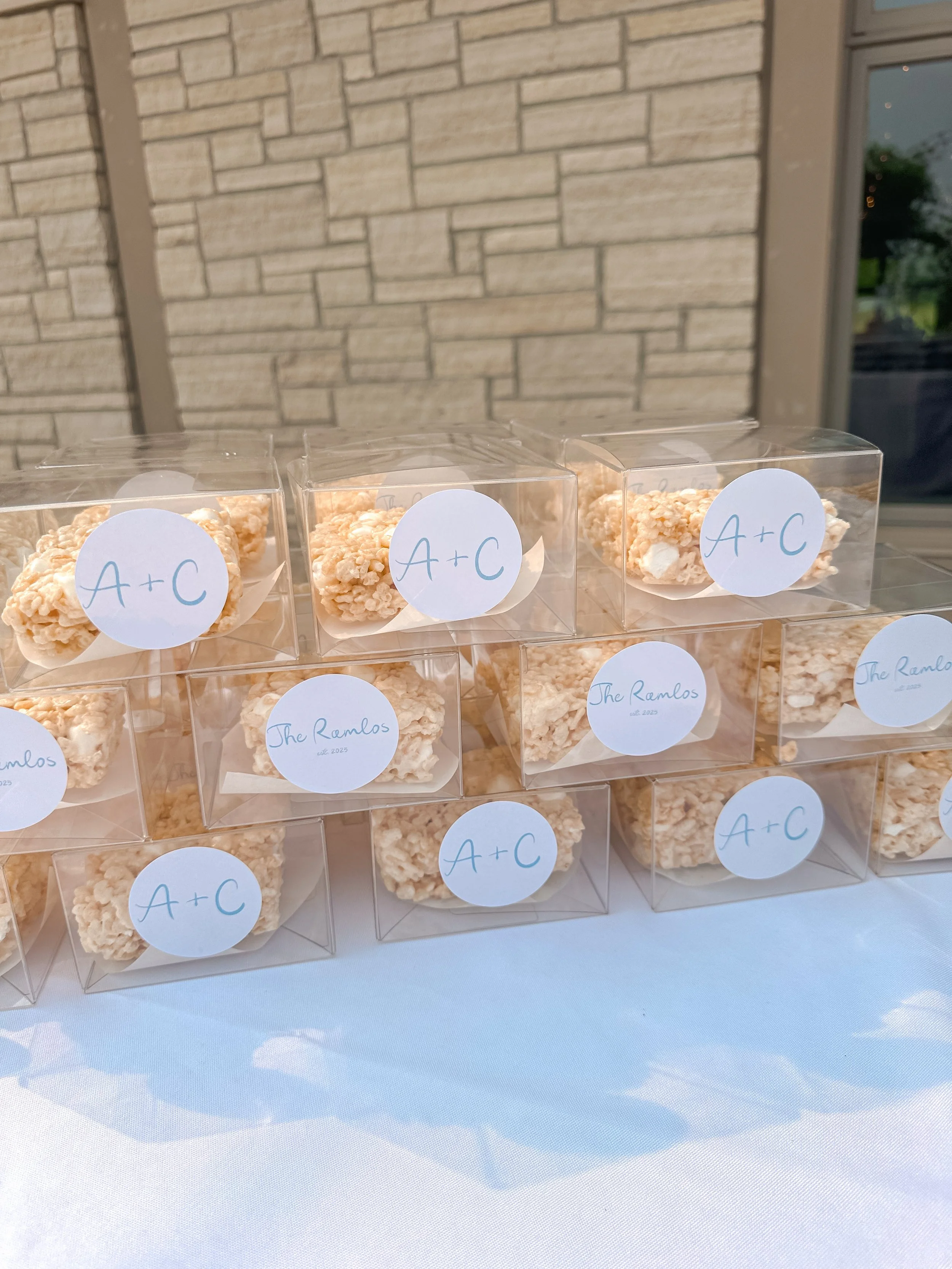 Clear plastic boxes with rice crispy treats labeled 'A+C' and 'The Ramlos' on a table.