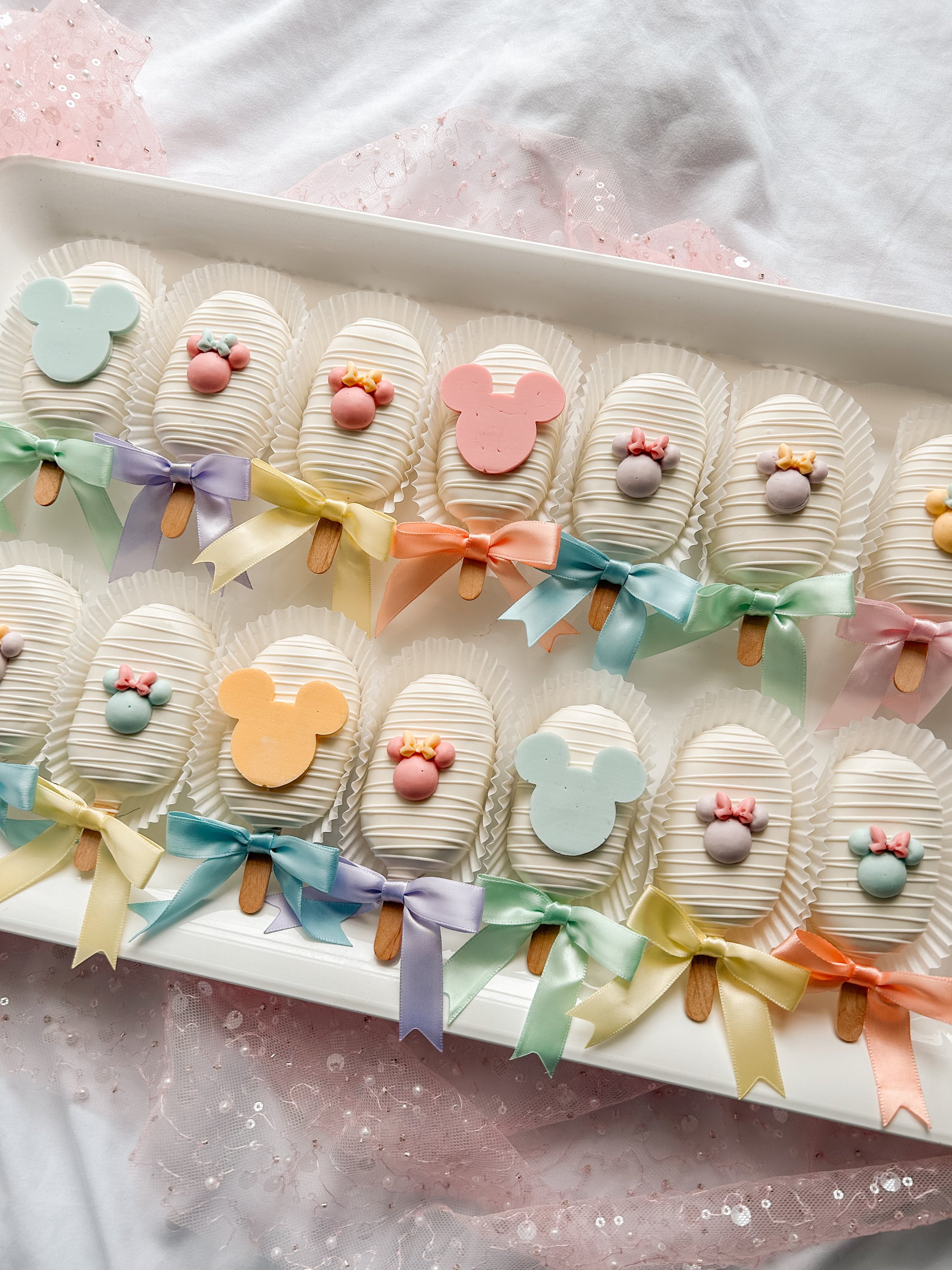 Assorted decorative cake pops with pastel Mickey Mouse and Minnie Mouse shapes and bows, arranged on a white tray with colored ribbons and pink tulle.