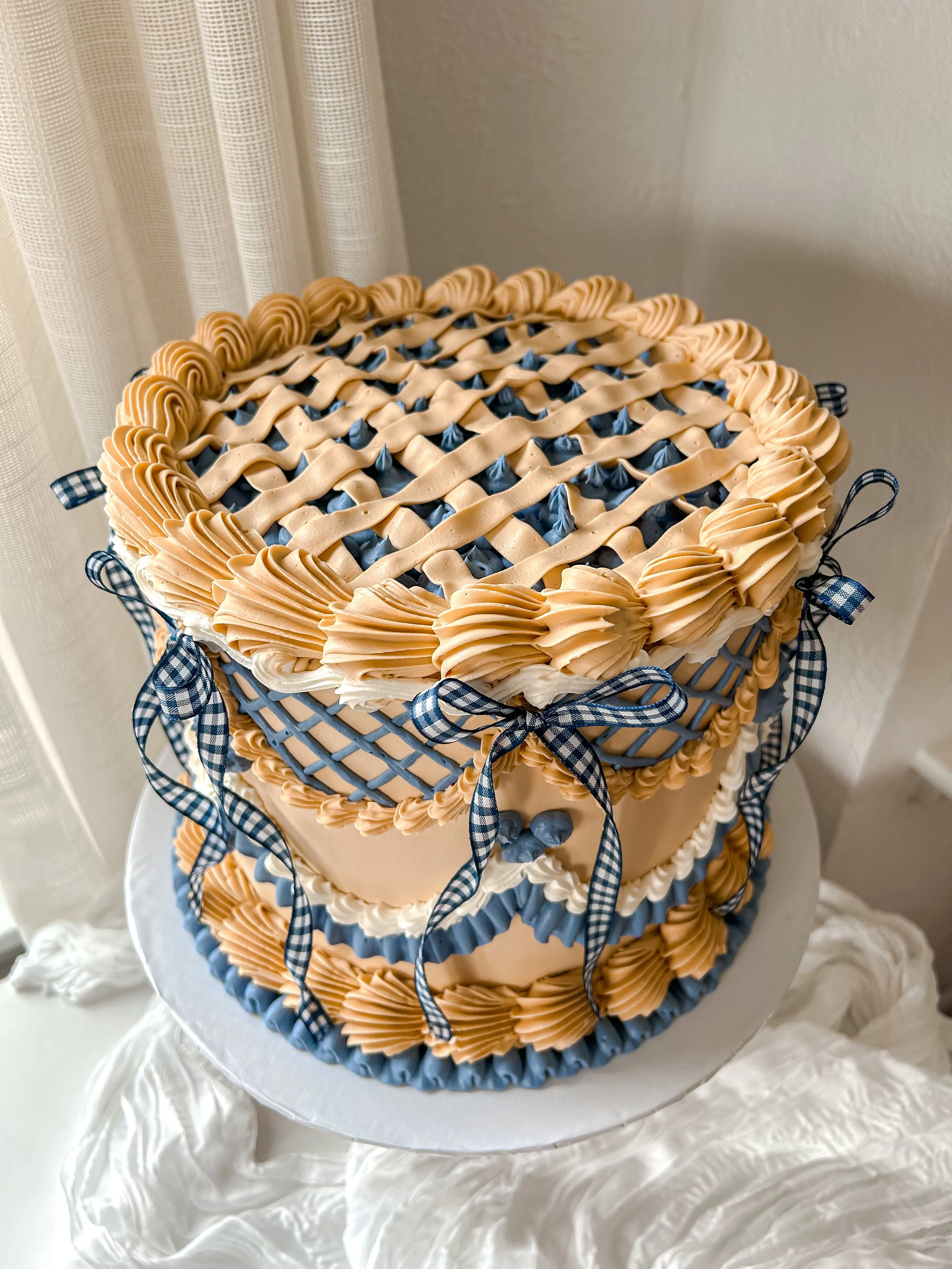 Decorative two-tiered cake with beige and blue icing, woven pattern design, topped with rosette decorations and tied with blue checkered ribbons.