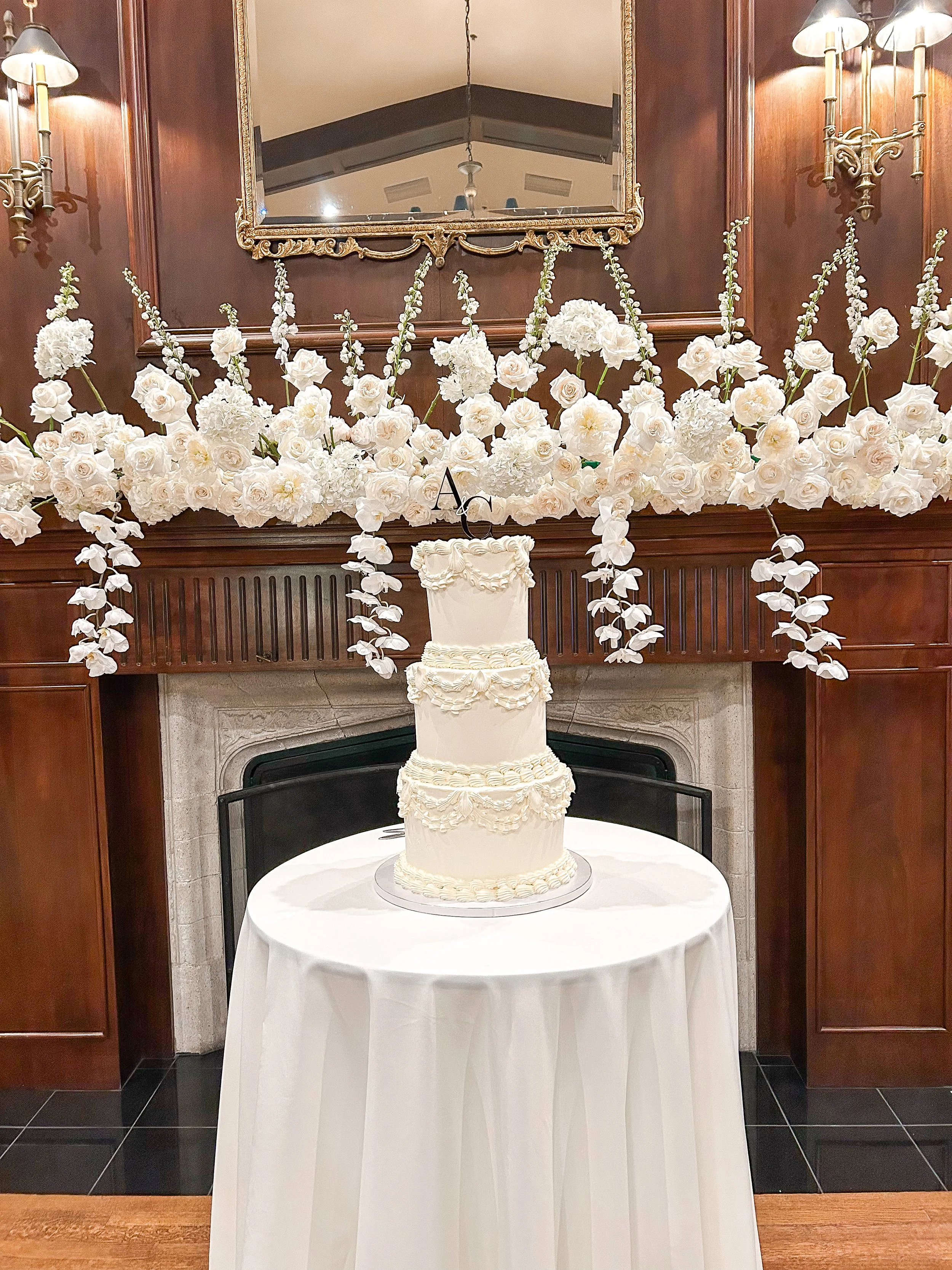 A multi-tiered white wedding cake with decorative icing, placed on a round table with a white tablecloth, in front of a decorated fireplace mantel with floral arrangements and a mirror.