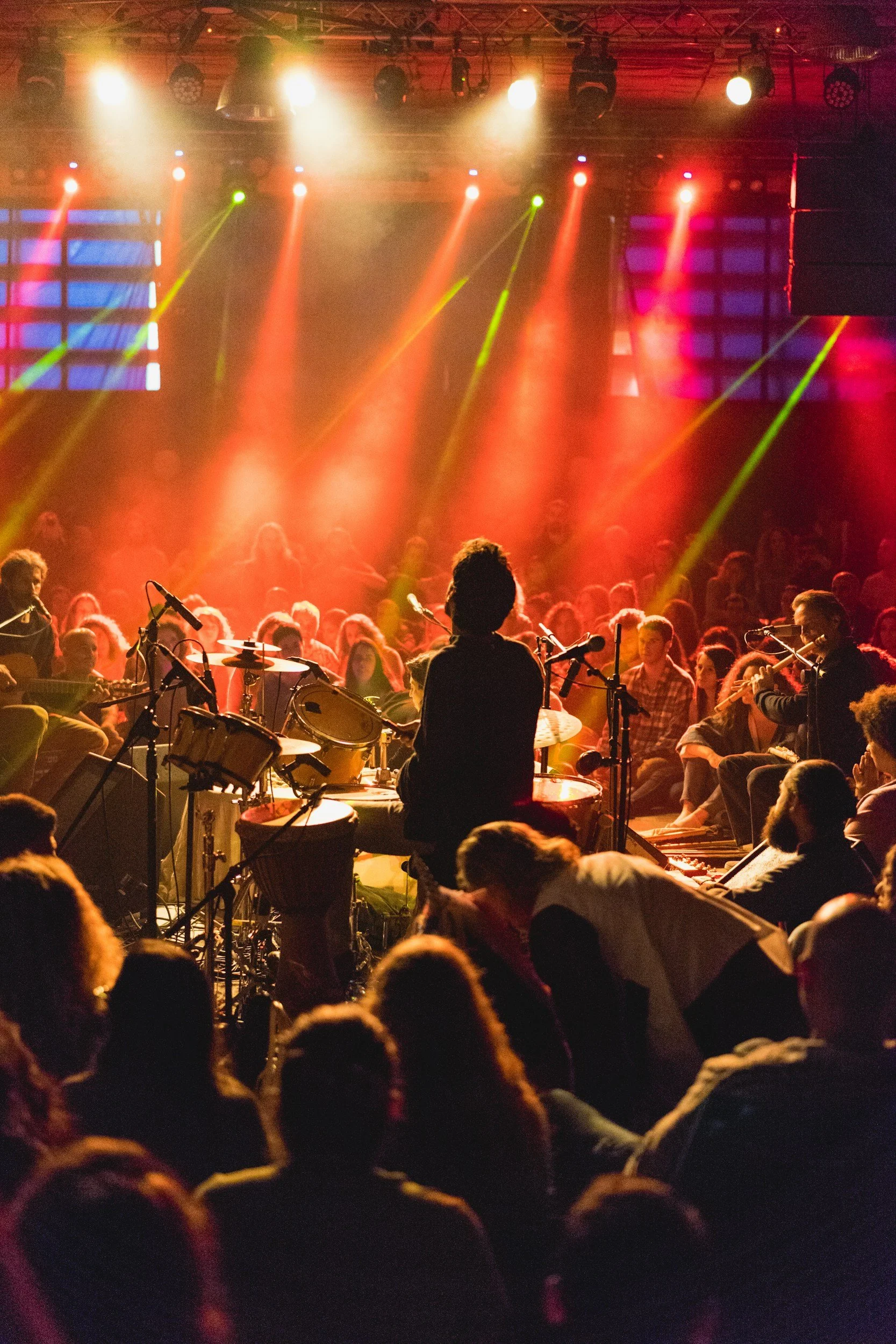 A live concert scene with a drummer performing on stage, surrounded by percussion instruments, with an audience in front. Bright stage lights in yellow, red, and green illuminate the scene and colorful LED screens are visible in the background.