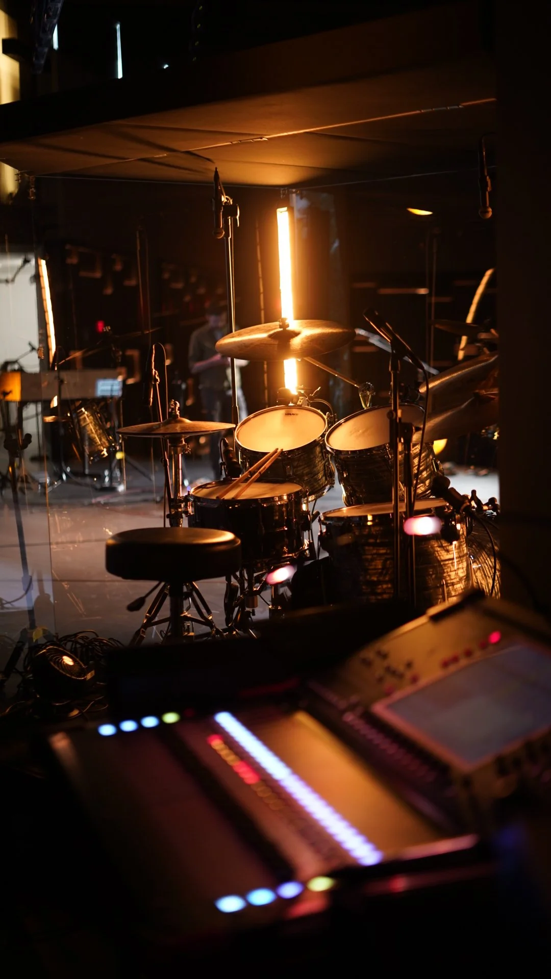 A drum set on stage illuminated by warm lighting, with a music mixer in the foreground.