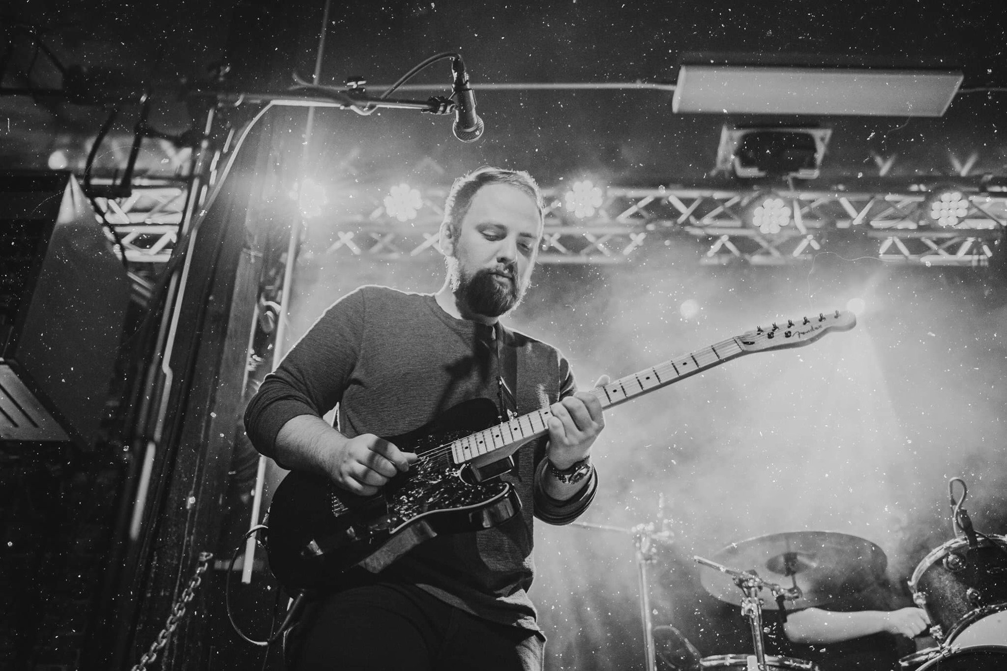 A black and white photo of Nathan Stumpf playing an electric guitar on stage, with stage lights and equipment in the background.