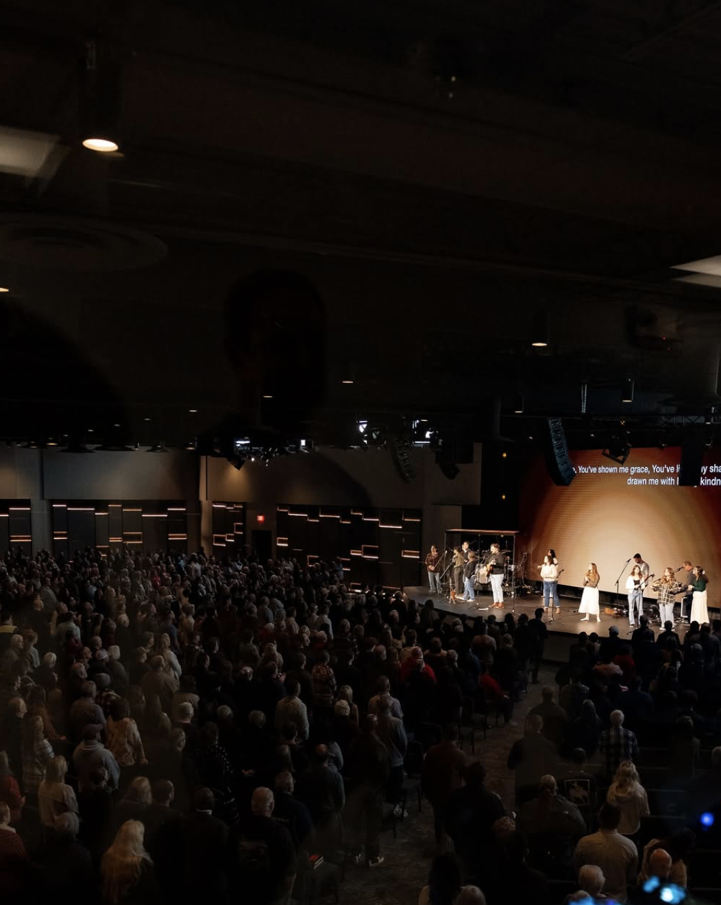Large indoor concert or worship event with a band and singers on stage, audience standing and facing the stage, with a screen displaying song lyrics in the background.