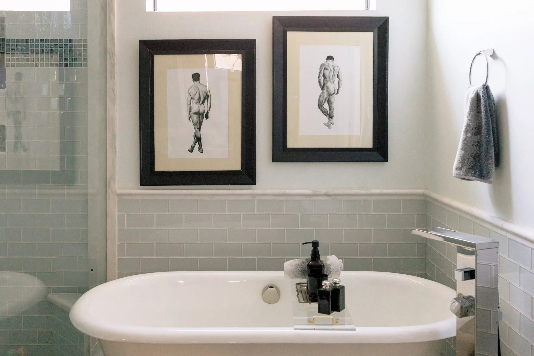 Bathroom with framed black-and-white sketches of male nudes on the wall above a white bathtub, with a wall-mounted towel ring holding a gray towel to the right.