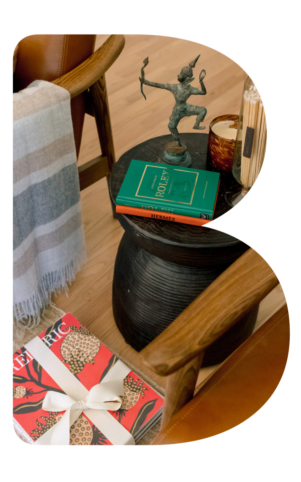 A cozy table arrangement with a decorative sculpture of a playful elf holding a bow, a turquoise book titled 'Pollex', a red book titled 'Hermès', and a wood container holding matches, set on a round black textured side table. Next to the table is a 
