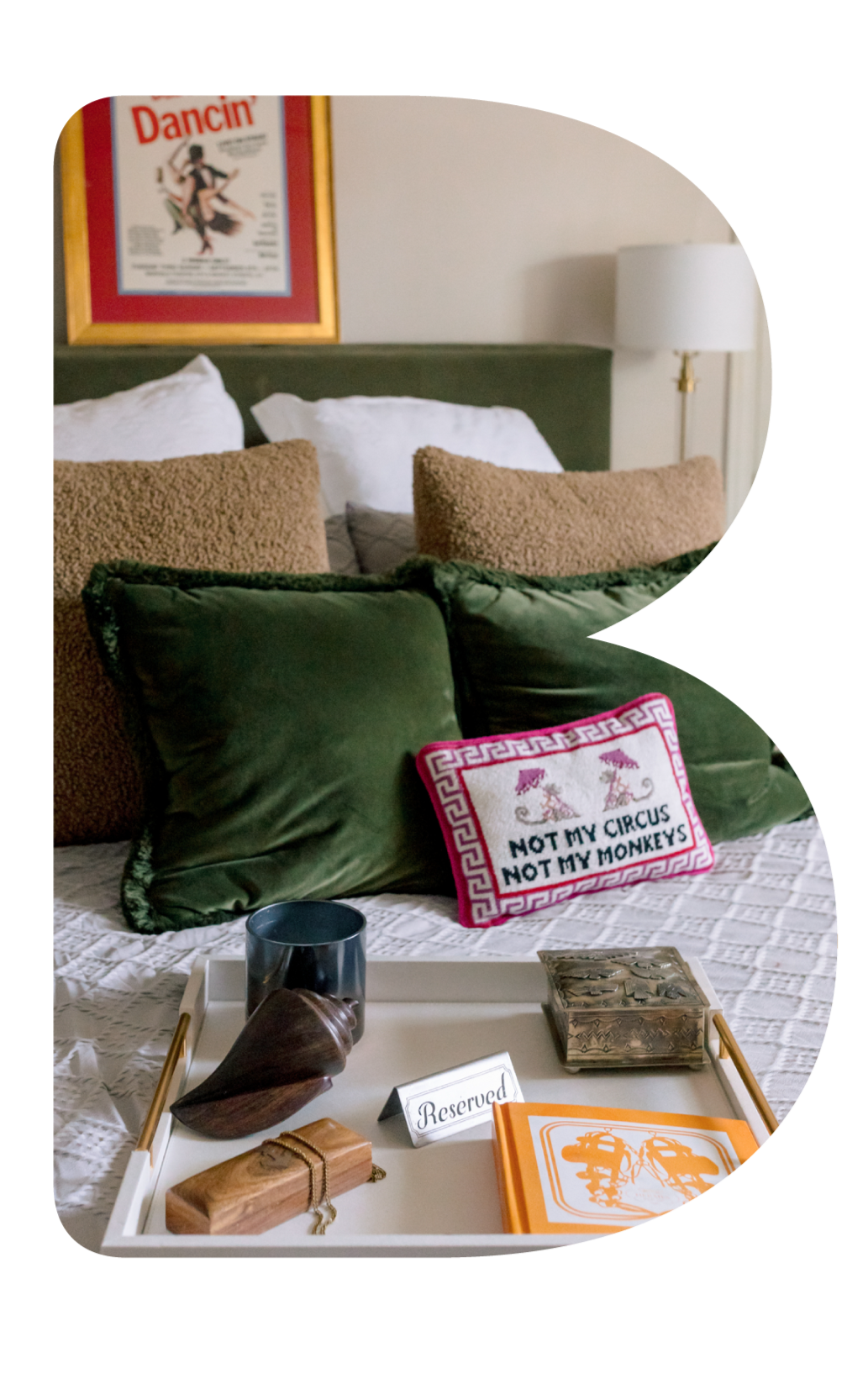 Close-up of a hotel bed with white linens, brown and green pillows, and a decorative pillow with the text 'Not My Circus Not My Monkeys'. A bedside lamp and framed poster are visible in the background, along with a tray on the bed holding a 'Reserved