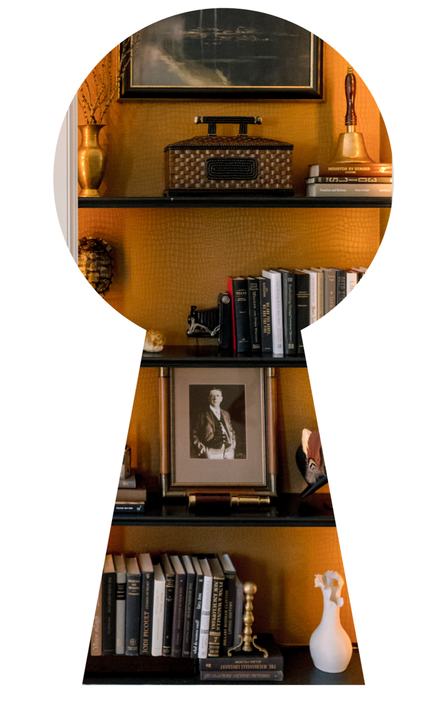 A wooden bookshelf with four shelves containing books, decorative items, framed photos, and art. The top shelf has a gold vase, a decorative box, and a brass bell. The second shelf has books and a small decorative object. The third shelf features a b
