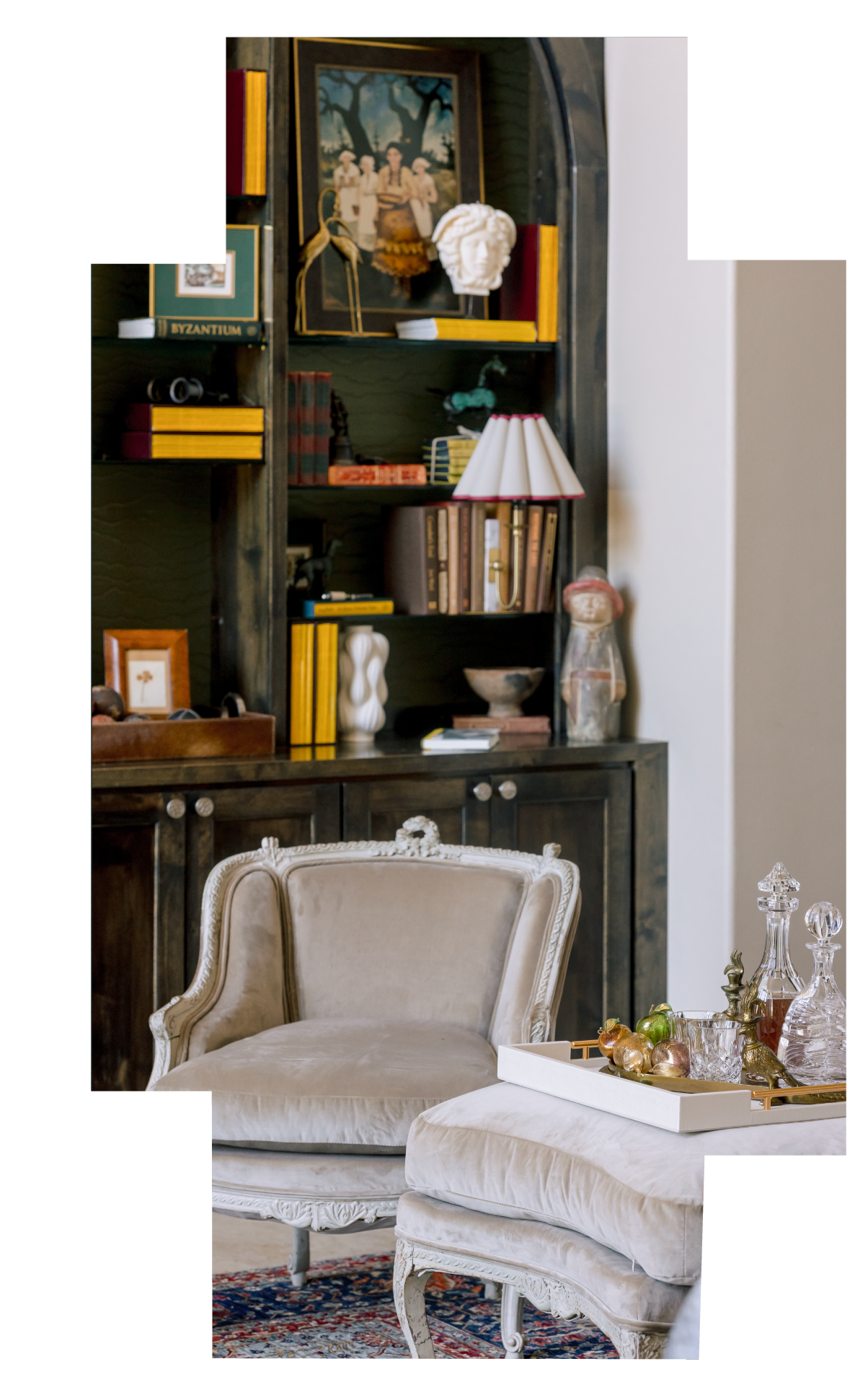 A living room with an ornate beige armchair, a vintage wooden bookcase filled with books and decorative items, a tray with glass decanters and ornaments on a white table.