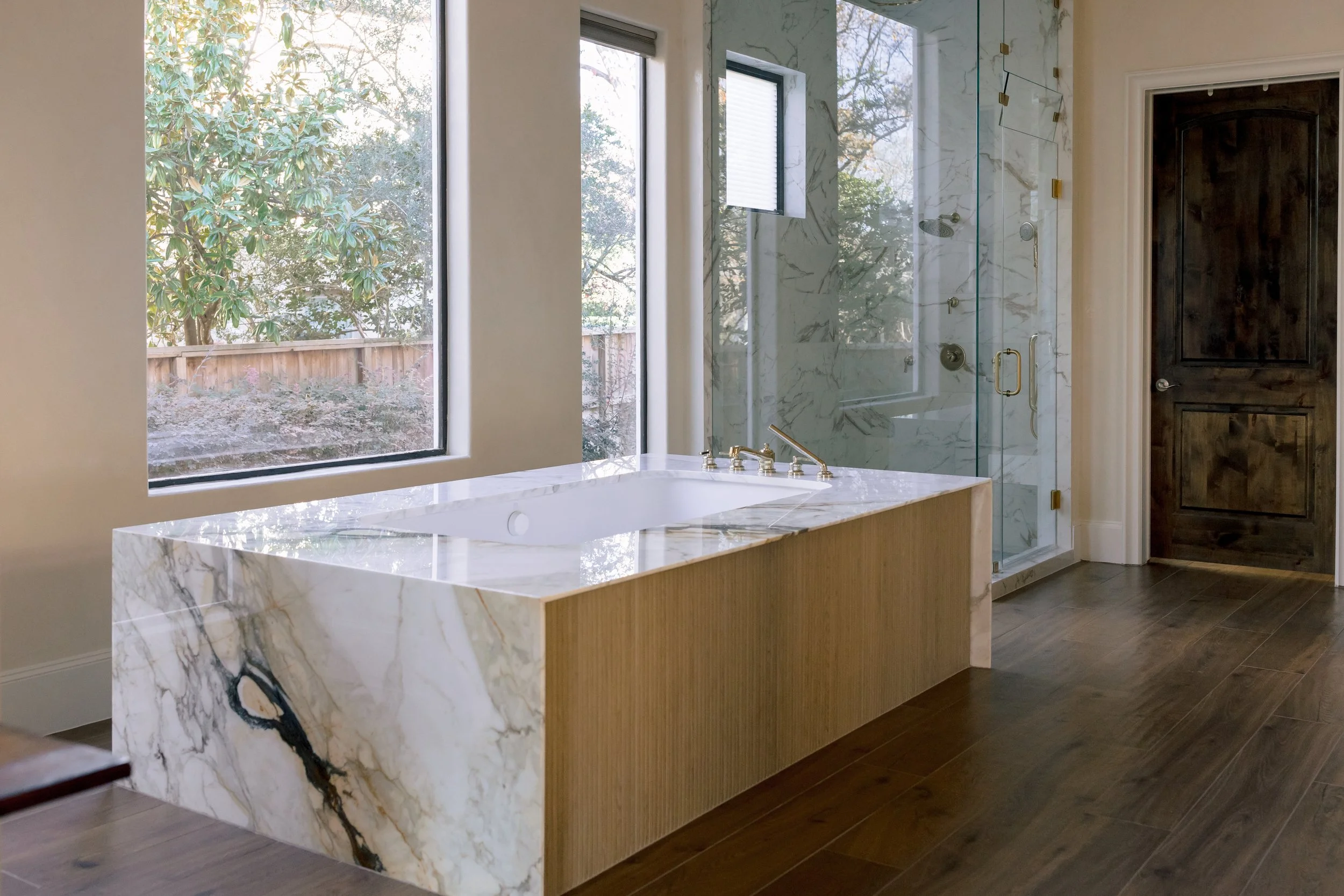 Modern bathroom with a whirlpool bathtub, large windows, a marble and wood vanity, and a glass-enclosed shower with marble walls and gold fixtures.