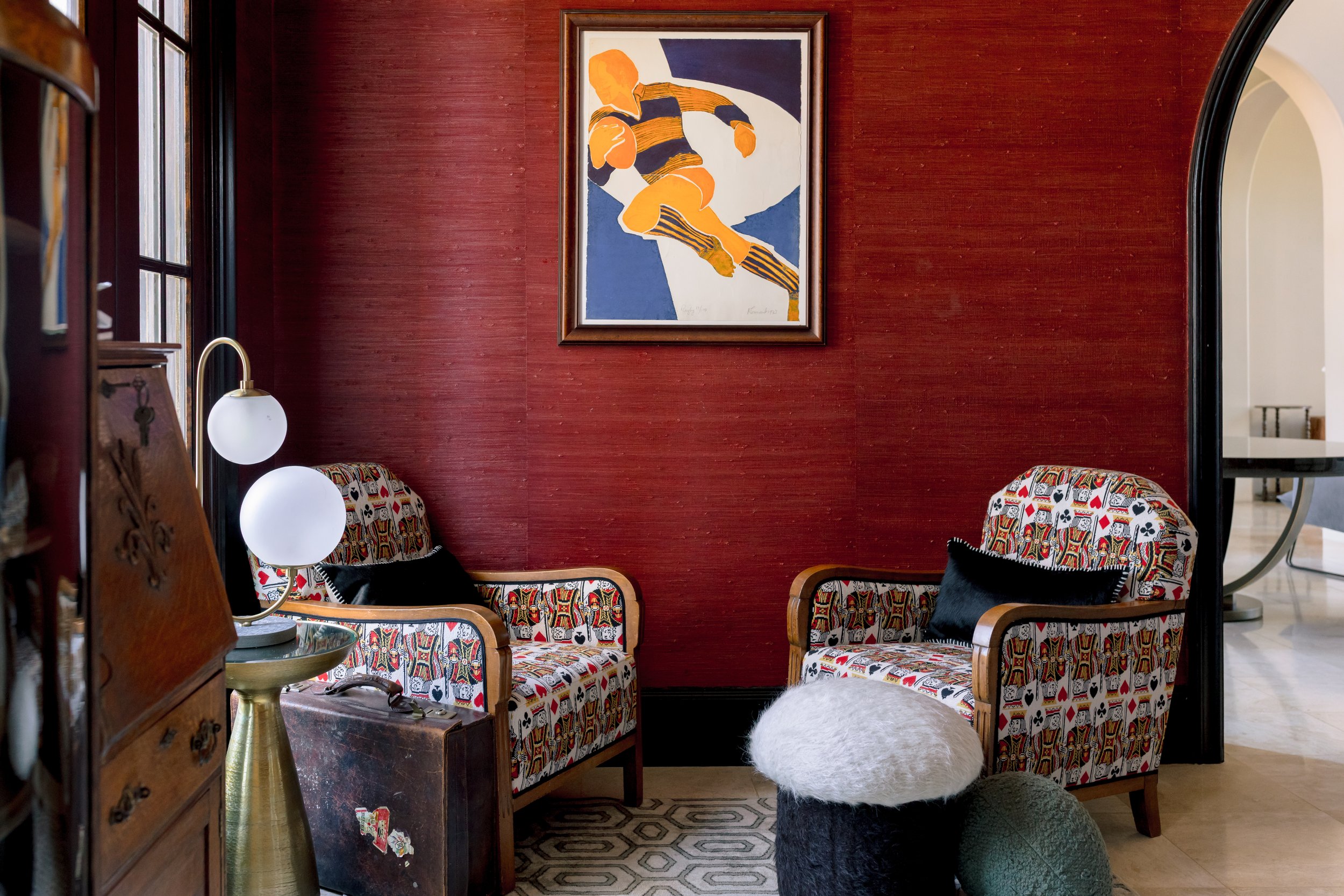 a cozy sitting area with two patterned armchairs, a black pillow, a furry ottoman, and a standing lamp with three round light bulbs. The space features a red textured wall, a framed abstract artwork, and is lit by natural light coming from a nearby w