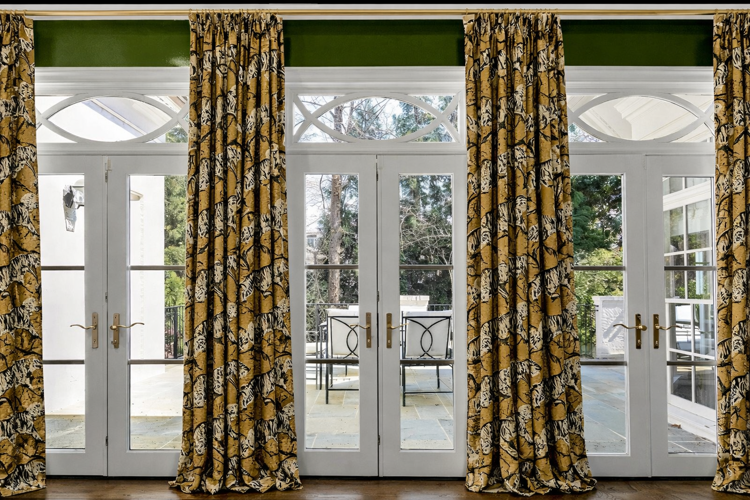 Interior view of French doors with glass panels, framed by yellow floral curtains, leading to an outdoor patio with chairs and surrounded by trees.