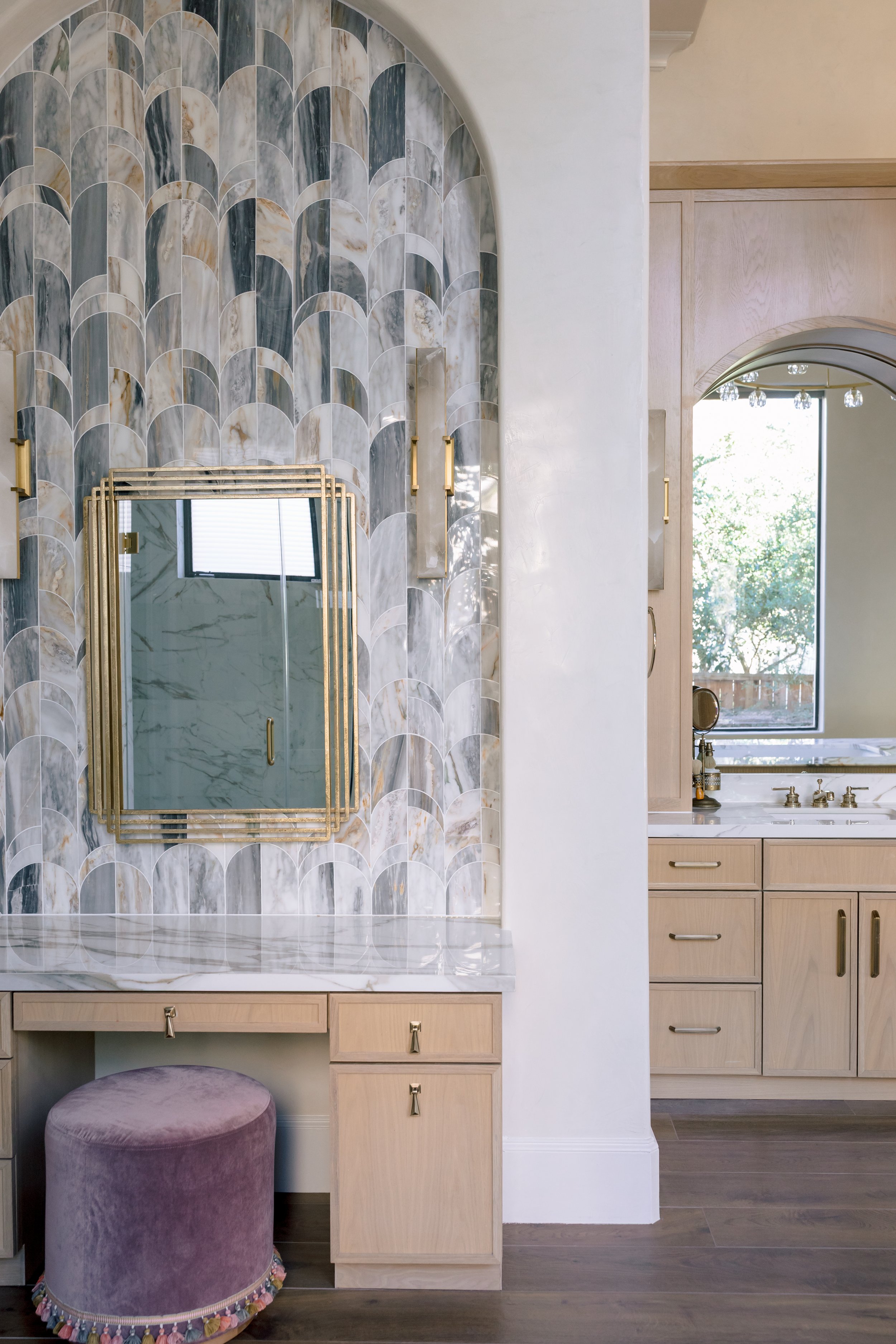 A vanity area with a marble countertop, a mirror with gold trim, light wood cabinets, and a purple velvet stool with colorful tassels at the bottom. In the background, a bathroom with a window, a sink, and gold fixtures is visible.