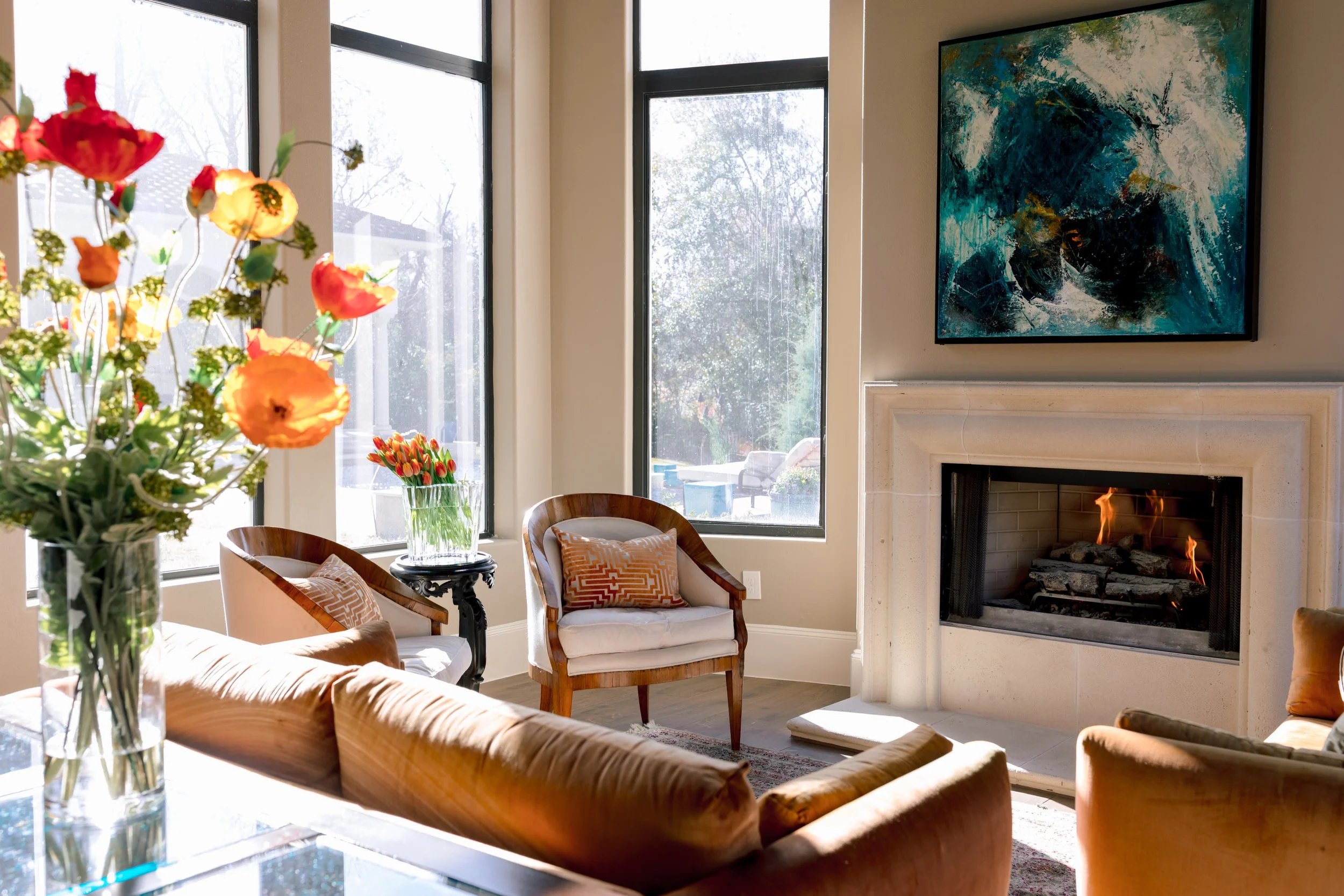 Living room with beige sofas, wooden armchairs with decorative pillows, a glass table with a vase of colorful flowers, large windows letting in sunlight, a fireplace with a fire burning, and abstract blue artwork hanging above the fireplace.