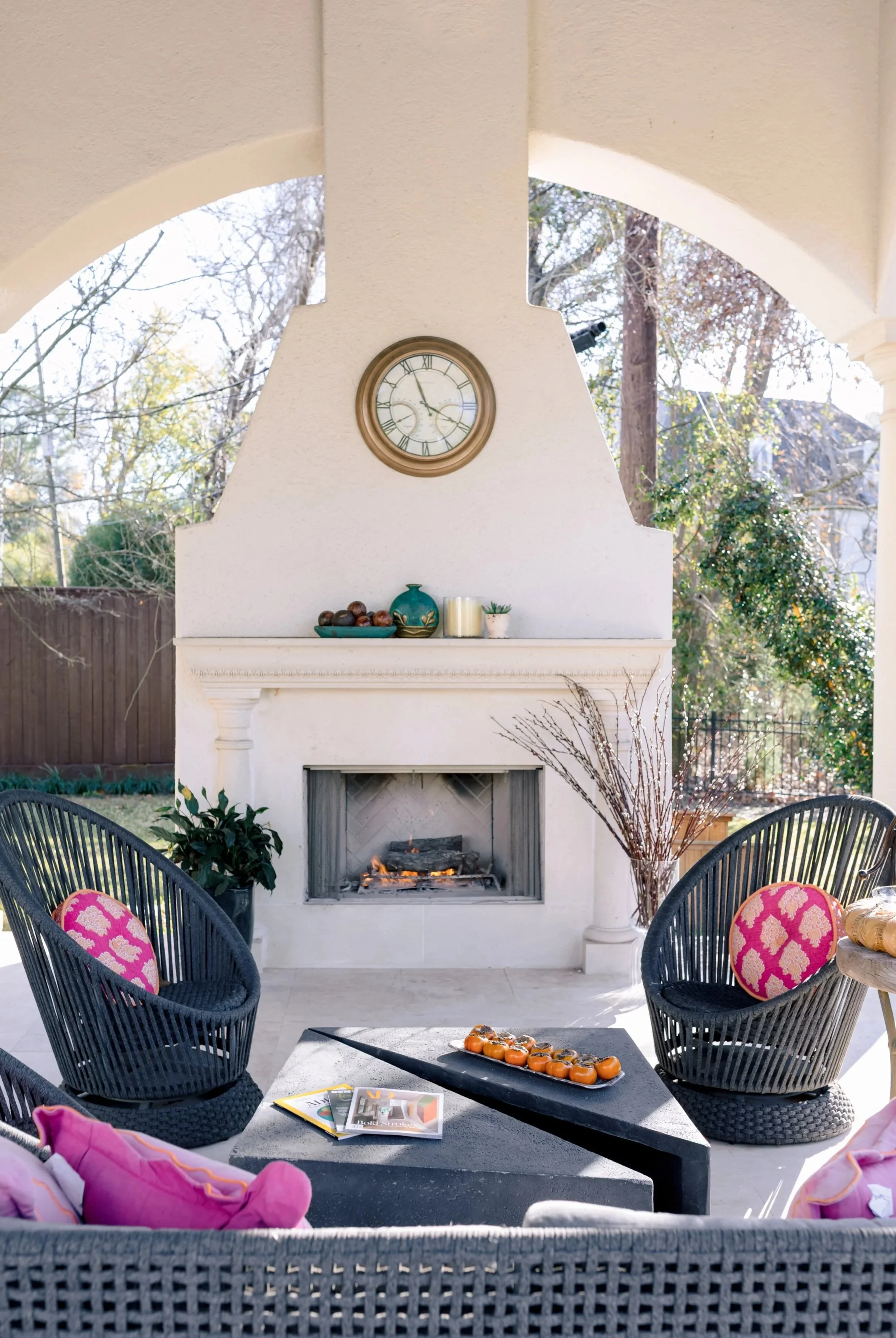 Outdoor patio with a white fireplace, two black chairs with colorful cushions, a coffee table with magazines and a tray of tangerines, and outdoor decor, under a covered archway with a clock on the wall.