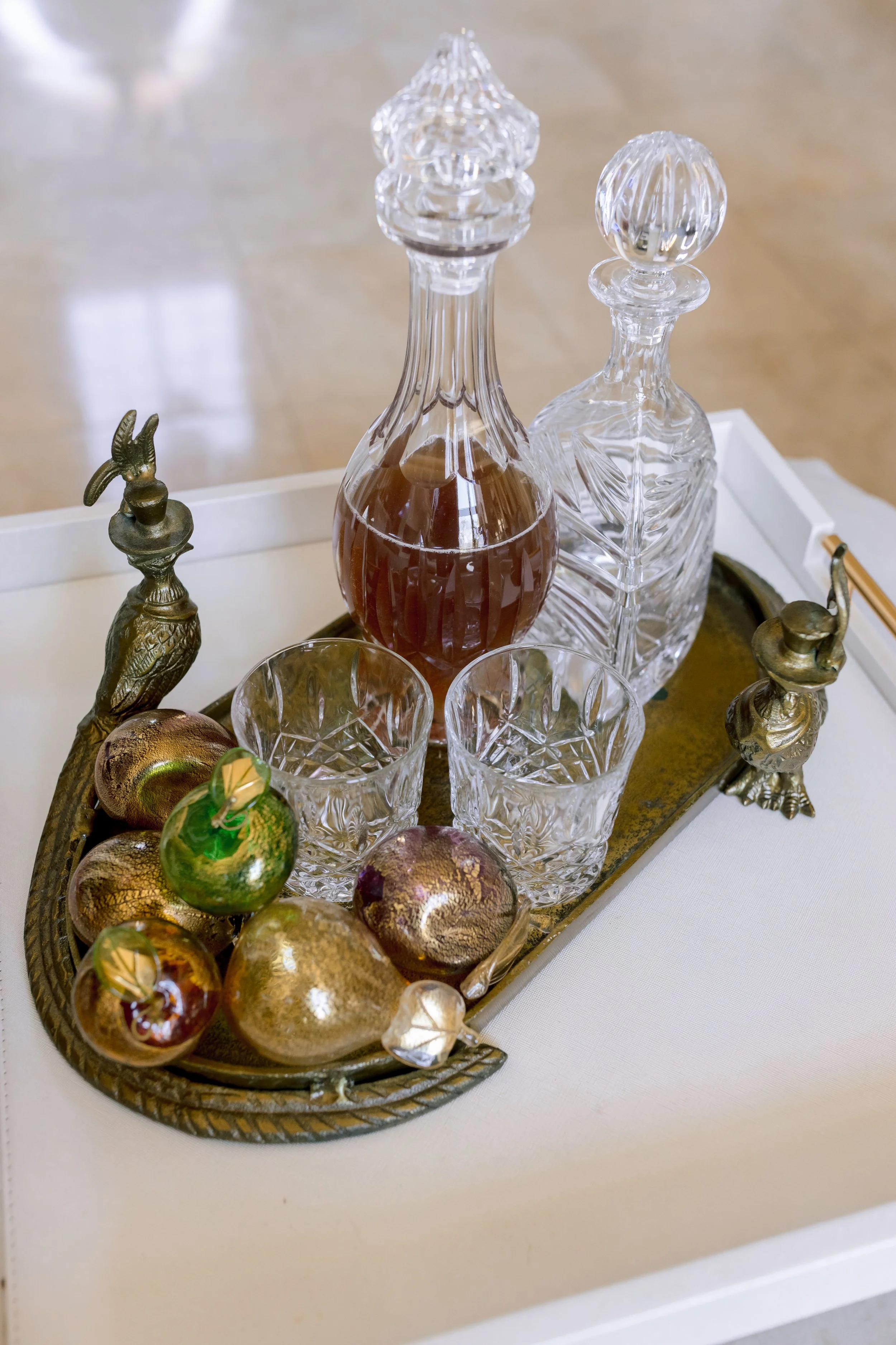 A tray with a decanter filled with amber-colored liquid, two clear glass decanters, two cut crystal glasses, and decorative gilded objects resembling fruits, all on a white surface.
