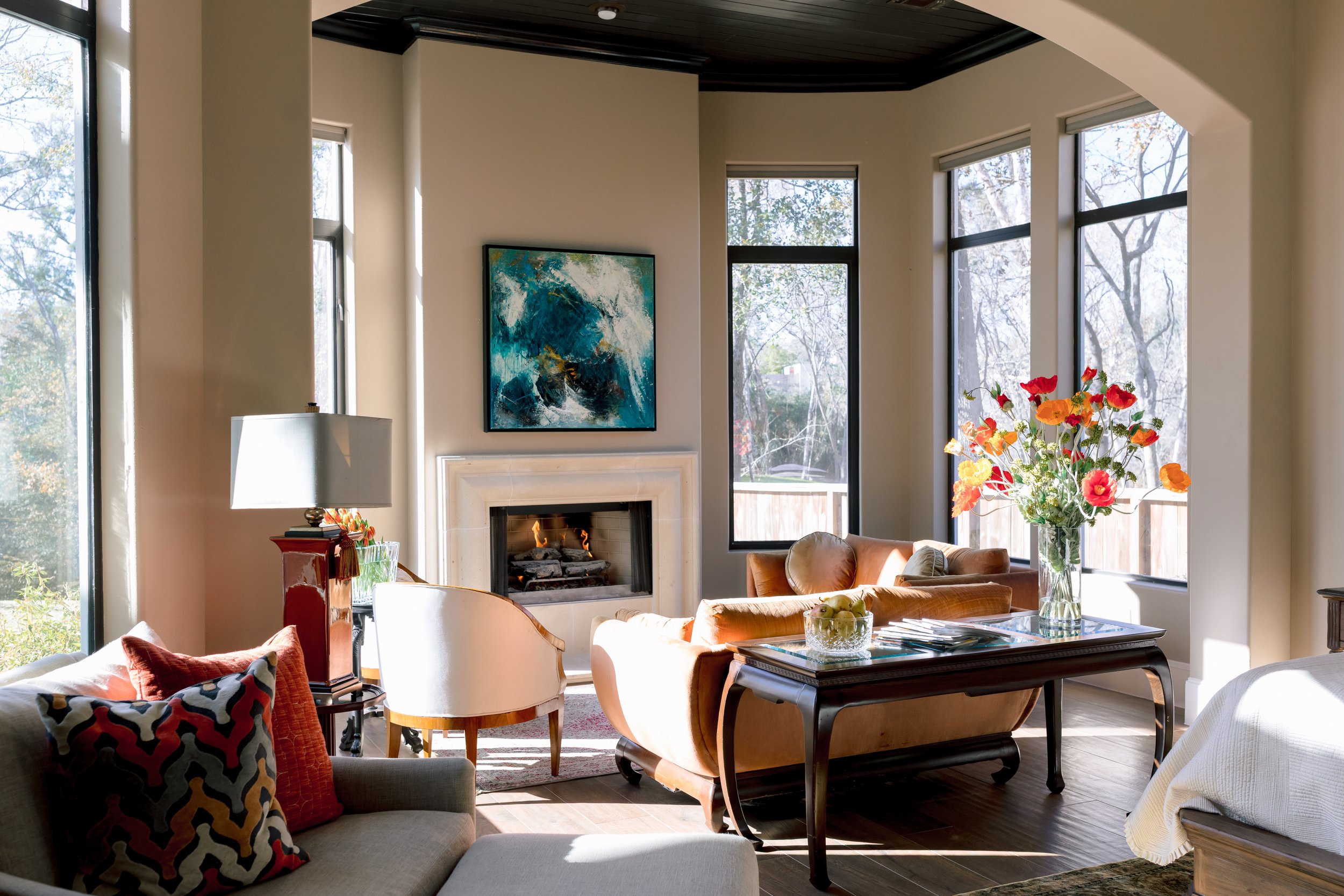 Living room with large windows, a fireplace, modern artwork, a beige sofa with colorful pillows, a table with a glass bowl of green apples, a vase with bright flowers, and a mix of contemporary and traditional furniture.