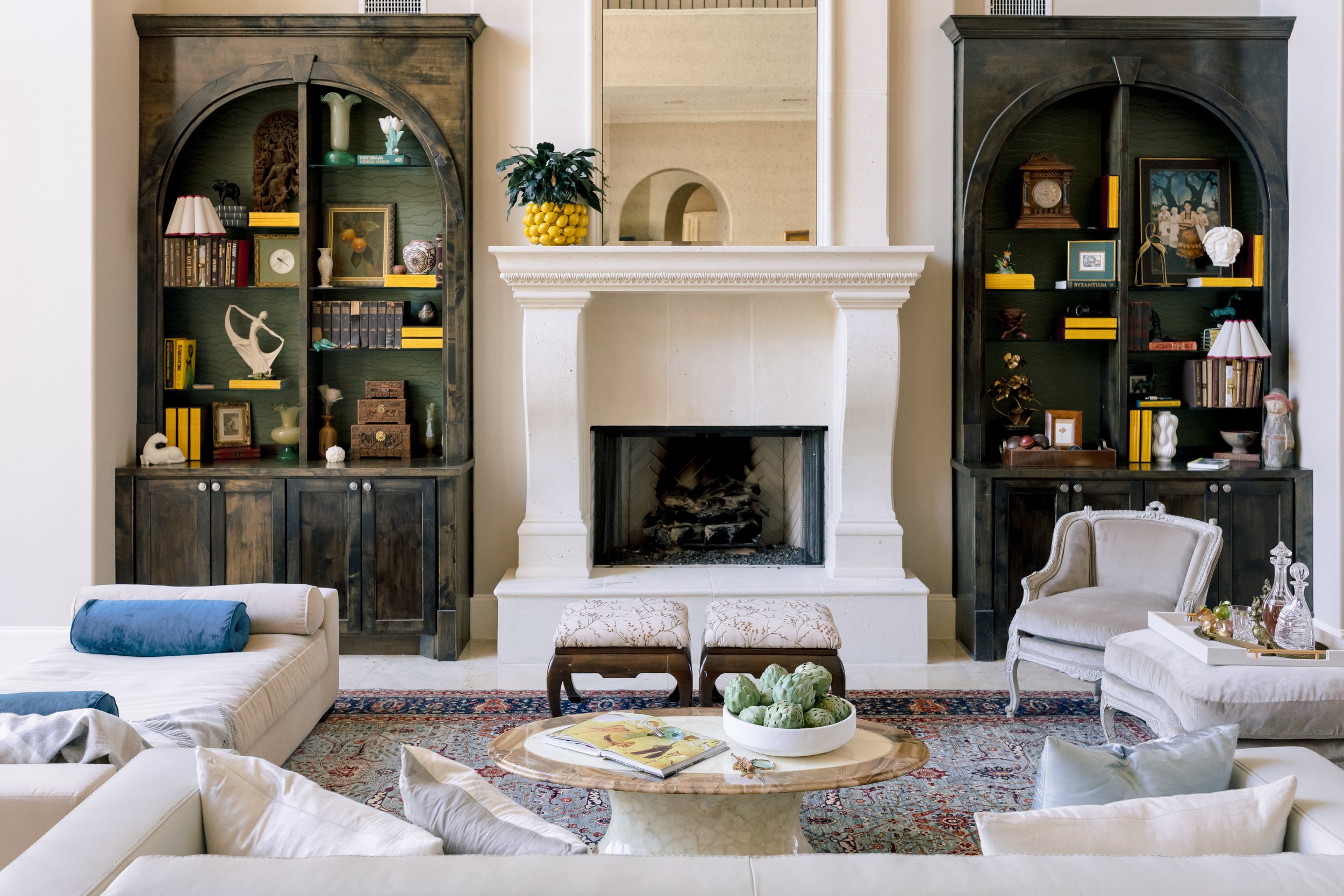 Living room with white fireplace, two dark wood bookshelves, cream sofas, a marble coffee table with a bowl of artichokes, and decorative items.