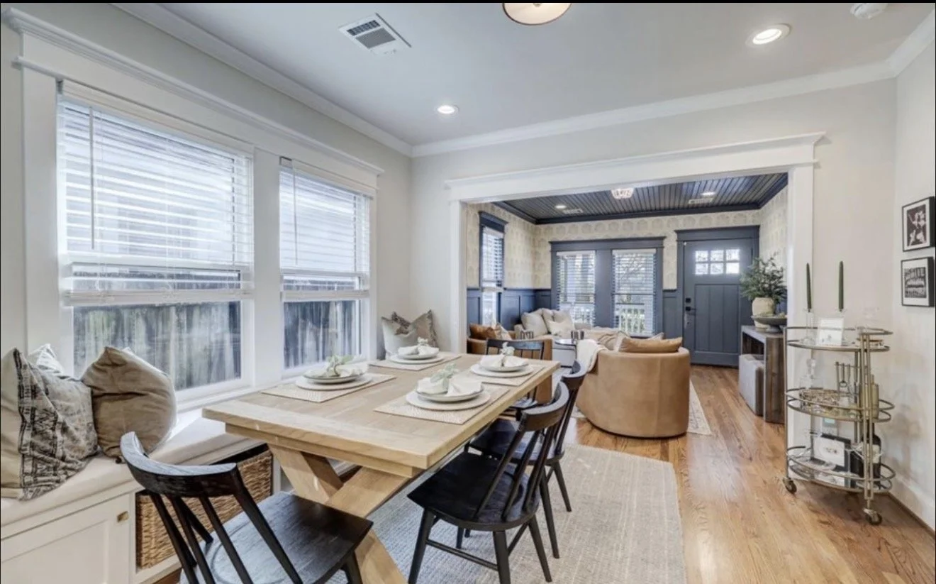 Open-concept living and dining room with large windows, white walls, wooden floors, a dining table with chairs, a sitting area with couches and armchairs, and a front door with windows and decorative trim.