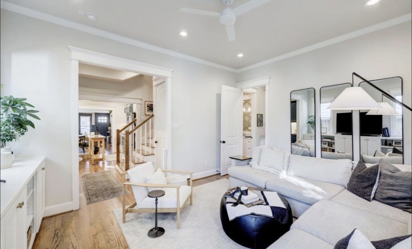 Bright living room with a white sectional sofa, black ottoman, and three geometric mirrors on the wall. There is a white ceiling fan, recessed lighting, and a doorway leading to a dining area and kitchen. A white sideboard with plants and decorative 