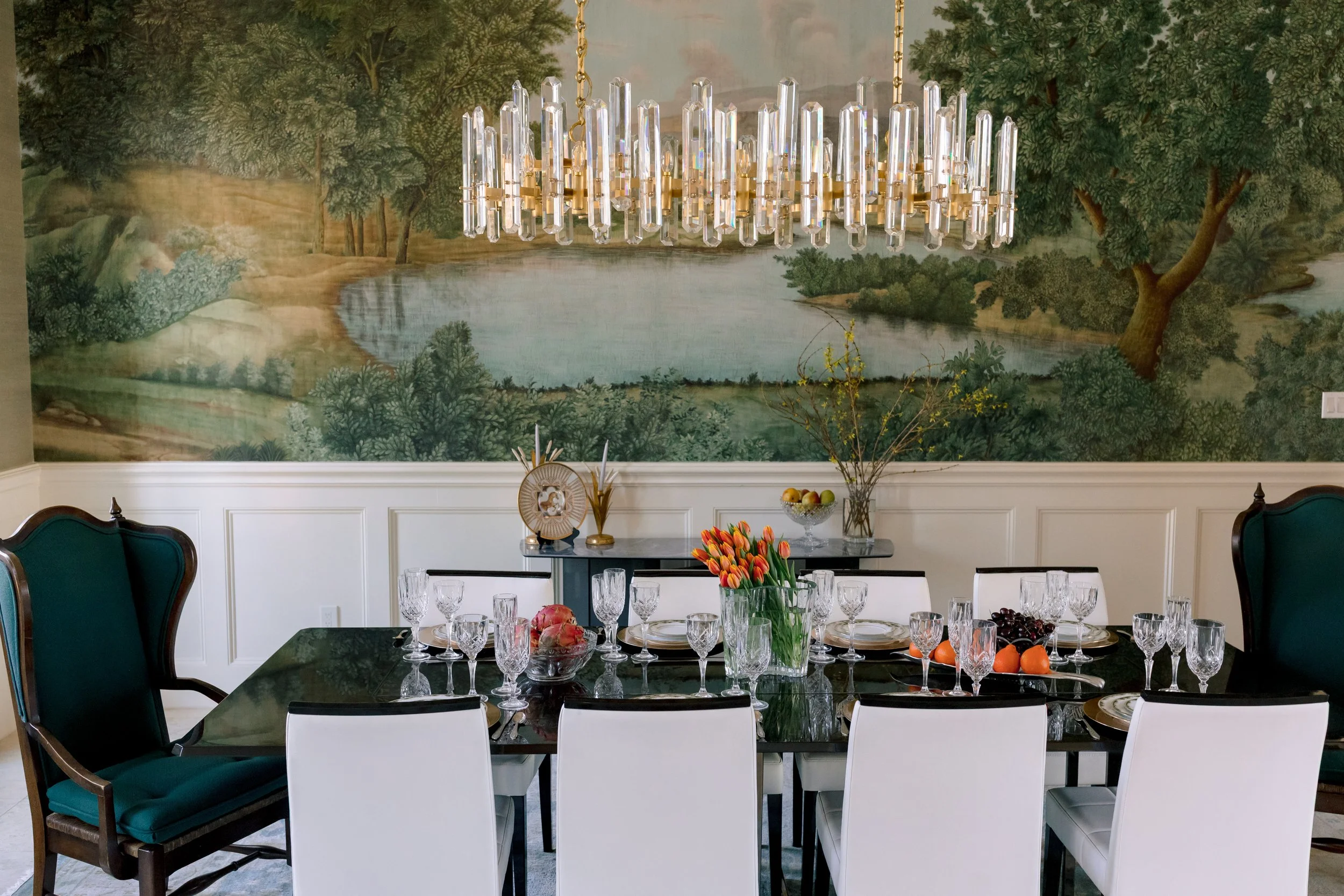A formal dining room with a black table set with crystal glasses and decorative plates, surrounded by white and dark green chairs. Above, a modern chandelier with multiple vertical glass rods hangs. The background features a landscape mural of a lake