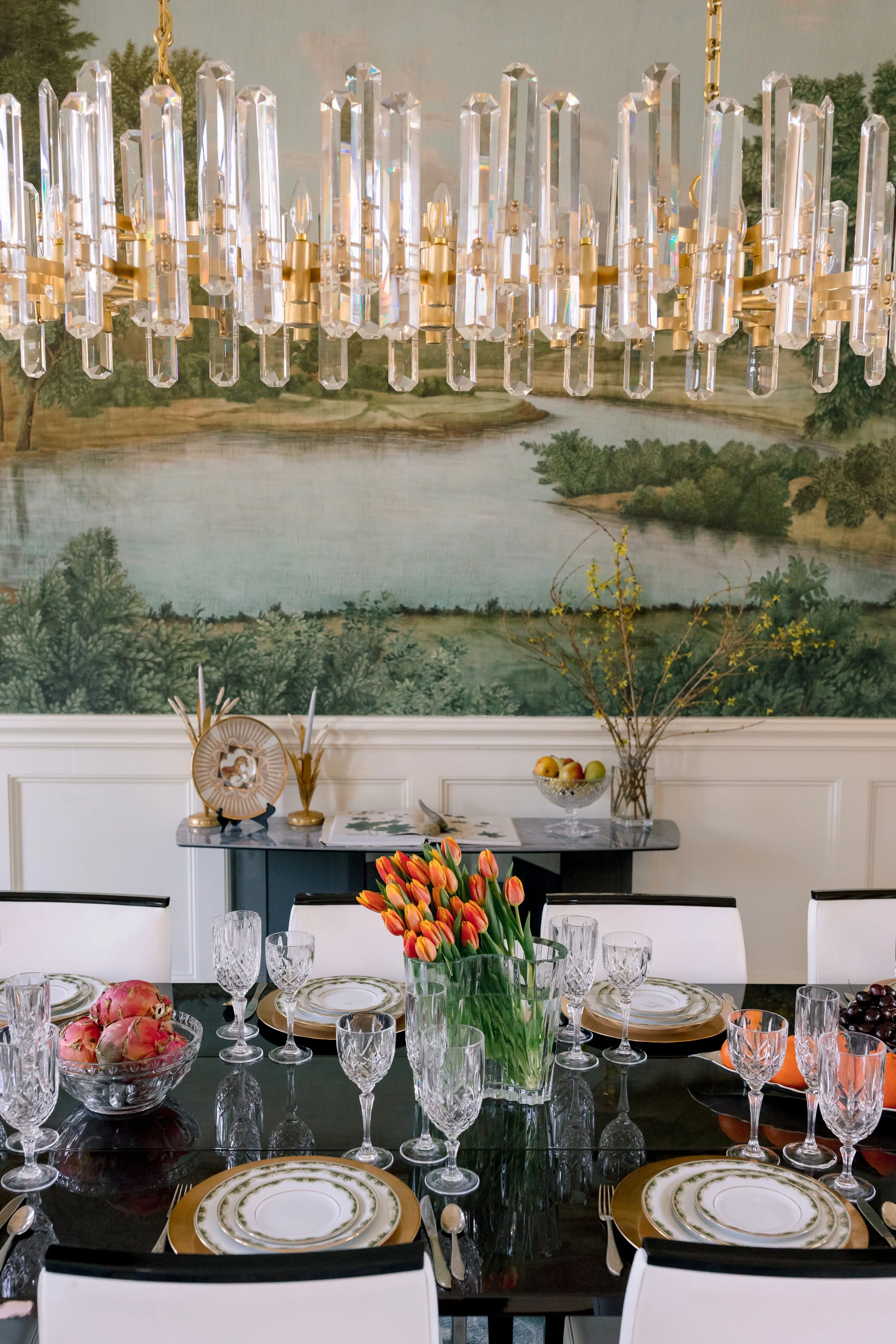 Formal dining room with a black table set with white and gold plates, multiple crystal glasses, and a large glass vase filled with orange tulips. The background features a landscape mural, a sideboard with decorative items, and a chandelier hanging a